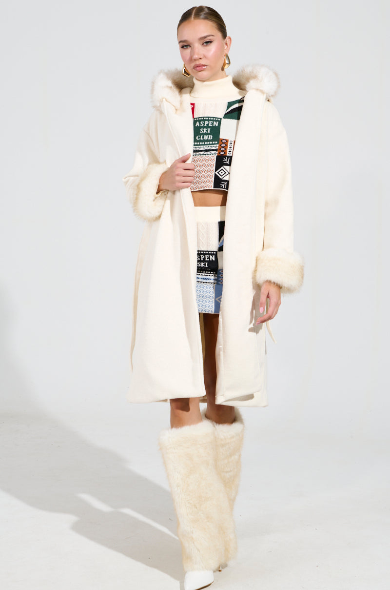 A woman is wearing the LILA FUR TRIM COAT, which features a white design with faux fur cuffs and a matching hood, paired with boots. Underneath, she wears a colorful patterned top and skirt. She stands against a plain white background, gazing ahead.
