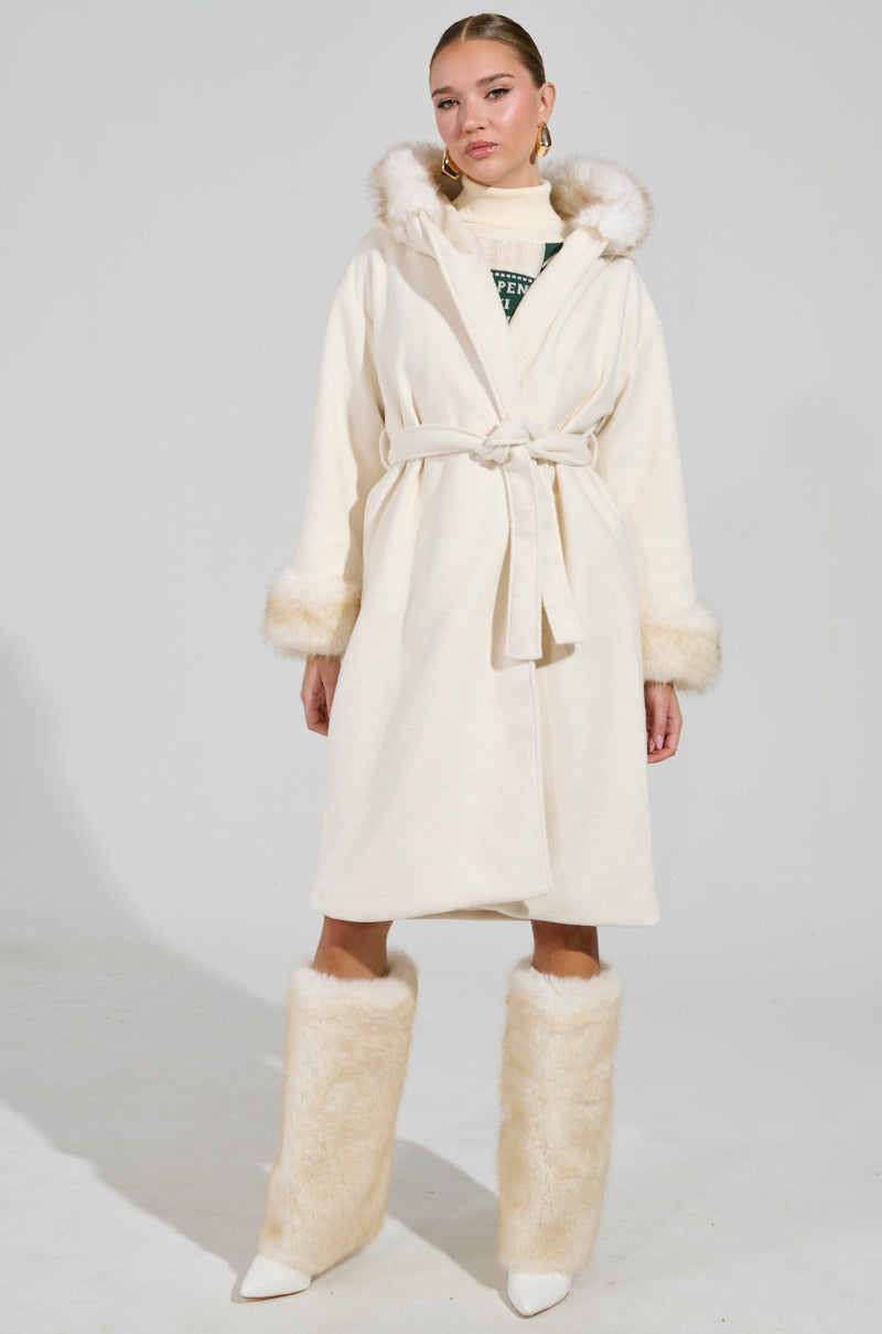 A person dressed in the LILA FUR TRIM COAT, featuring a cream color with a faux fur-lined hood and cuffs, a coordinating belt, and accompanied by fluffy knee-high boots. The background is plain gray.