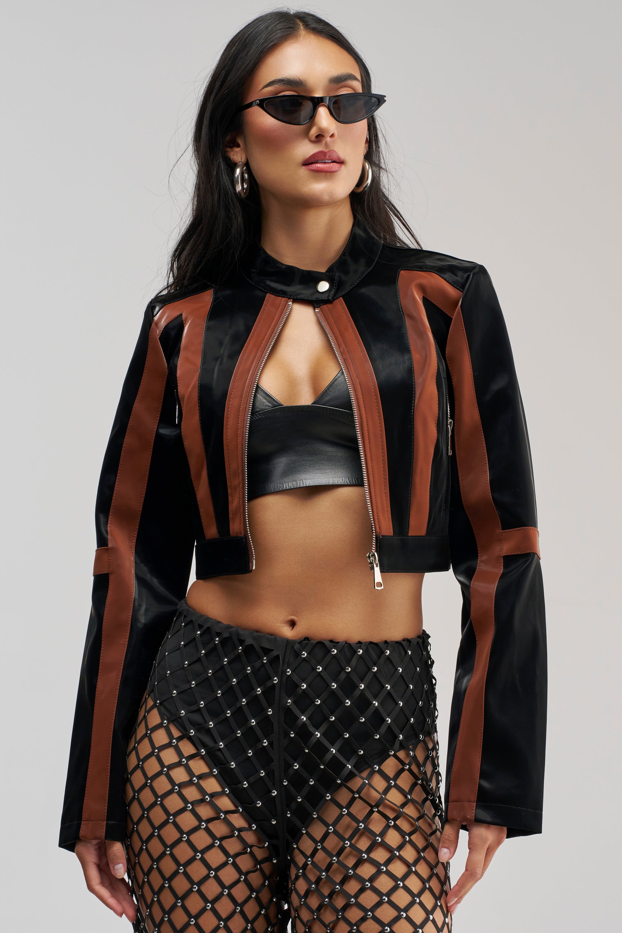 A woman poses confidently against a plain background, wearing the MADE YOU LOOK LIQUID VELVET RACER MOTO jacket over a black leather crop top, paired with black shorts and a fishnet skirt. She accessorizes her look with long hair, sunglasses, and hoop earrings.
