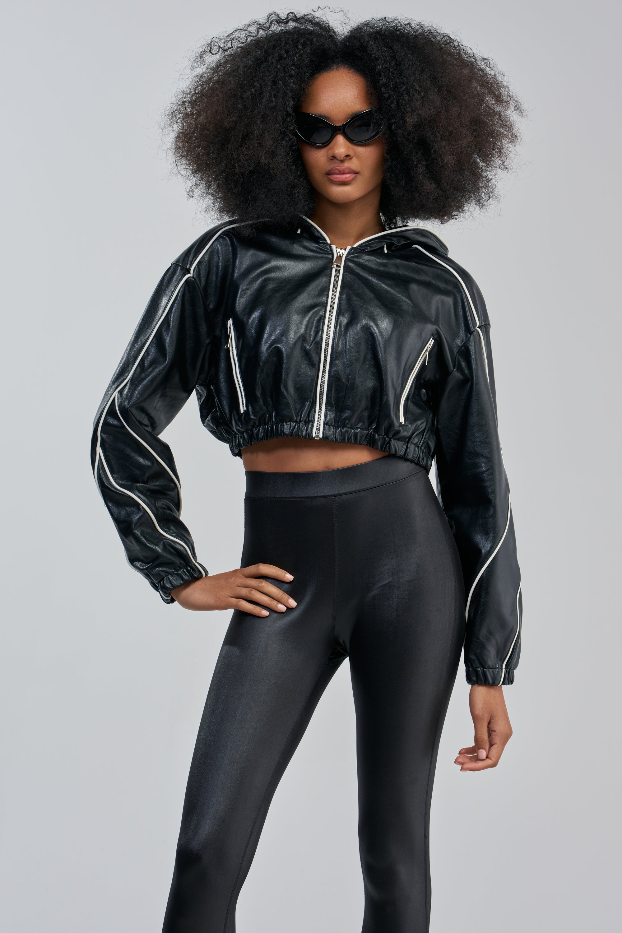 A woman wearing sunglasses, the FAUX LEATHER HOODED BOMBER in glossy black with white piping, and high-waisted black leggings stands confidently against a plain background.