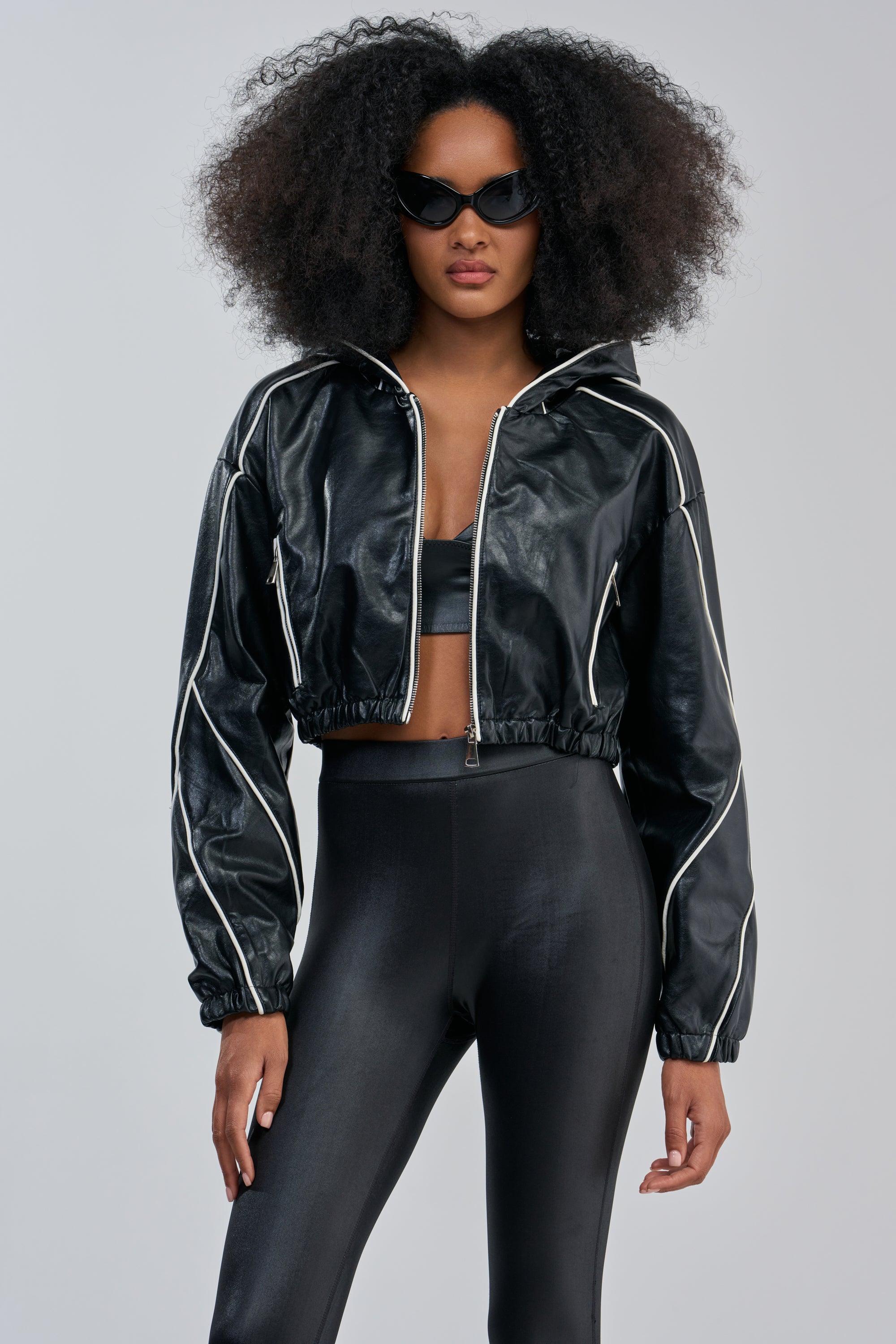 A person wearing black sunglasses and the FAUX LEATHER HOODED BOMBER—a glossy black faux leather cropped jacket with white piping—paired with matching high-waisted leggings, stands facing forward against a plain light gray background.
