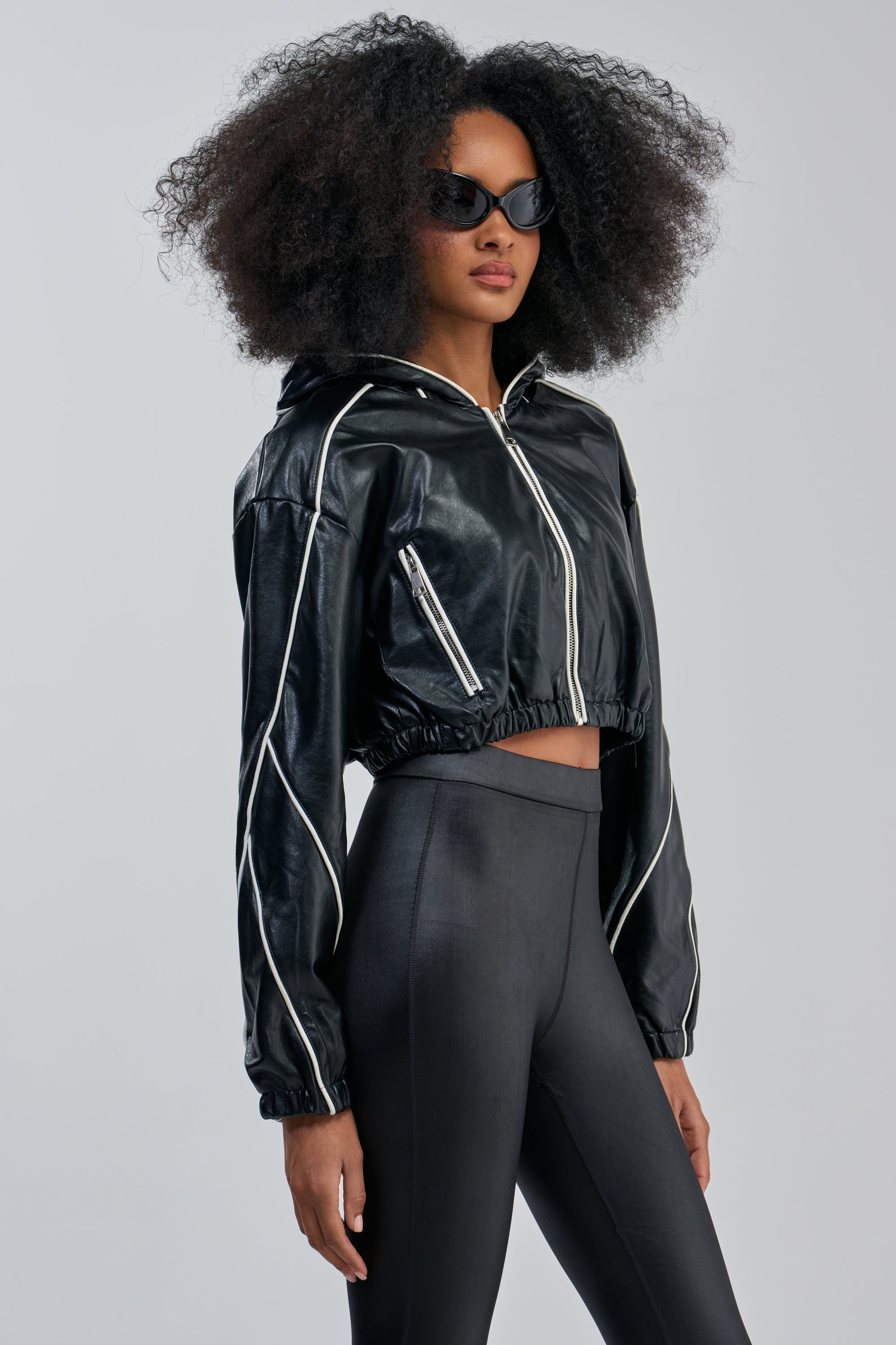 A woman with voluminous curly hair wears black sunglasses and the FAUX LEATHER HOODED BOMBER—a glossy black faux leather jacket with white piping—paired with high-waisted black leggings, standing against a plain light background.