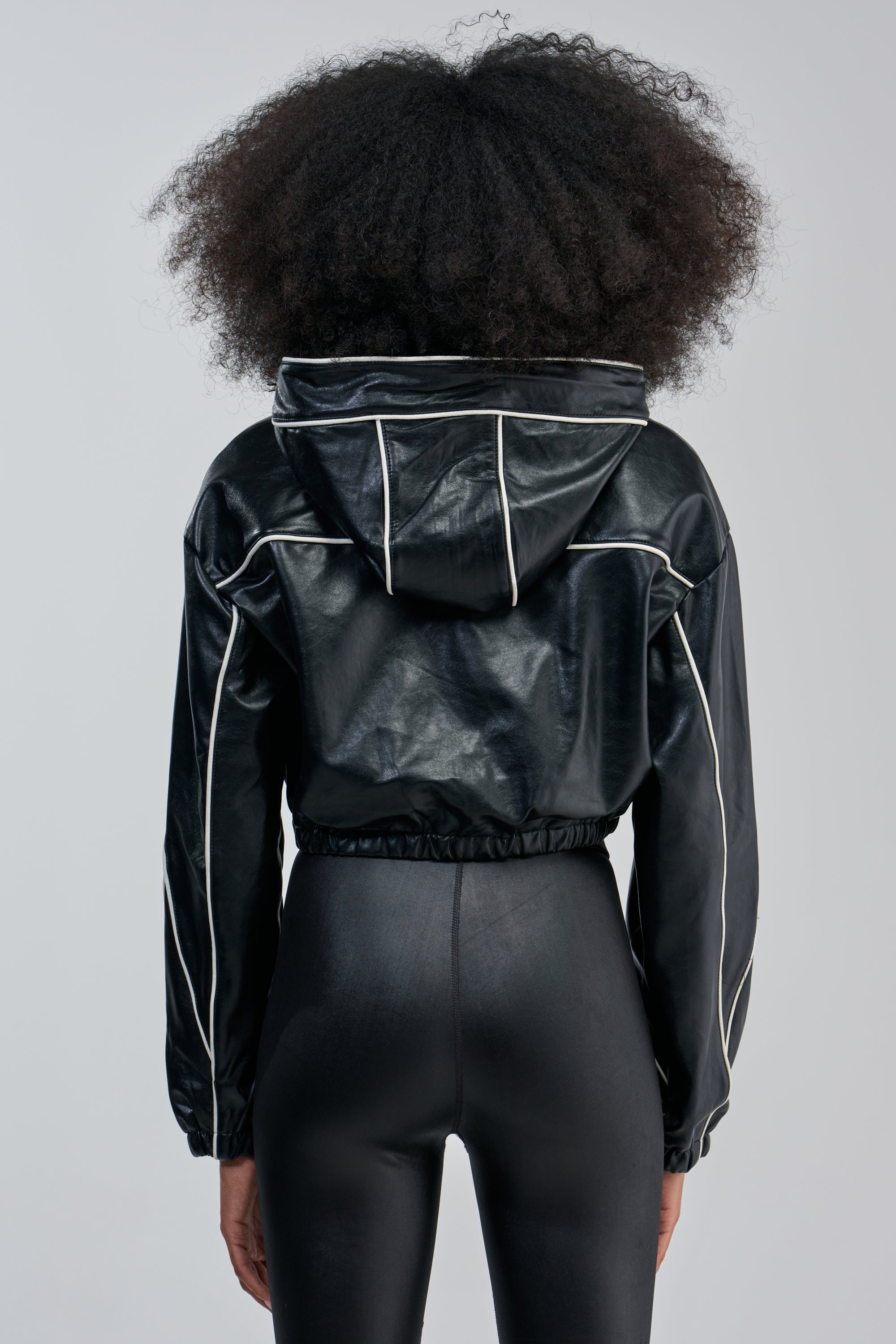 A person with curly hair models the FAUX LEATHER HOODED BOMBER in glossy black—with white piping—paired with matching black pants, shown from behind against a plain light background.
