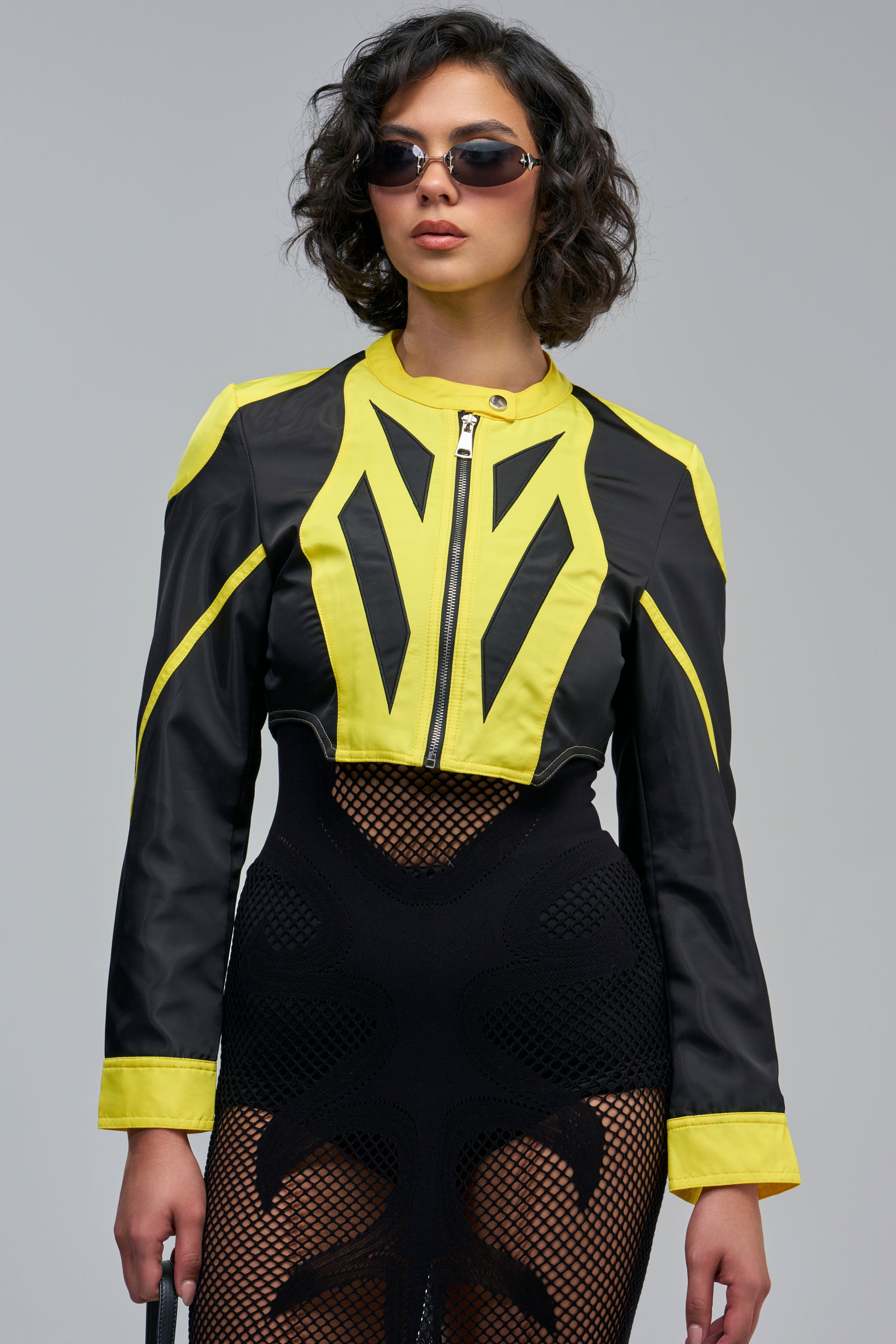 A cropped black and yellow FLOOR IT NYLON RACER MOTO jacket worn over a black mesh bodysuit.