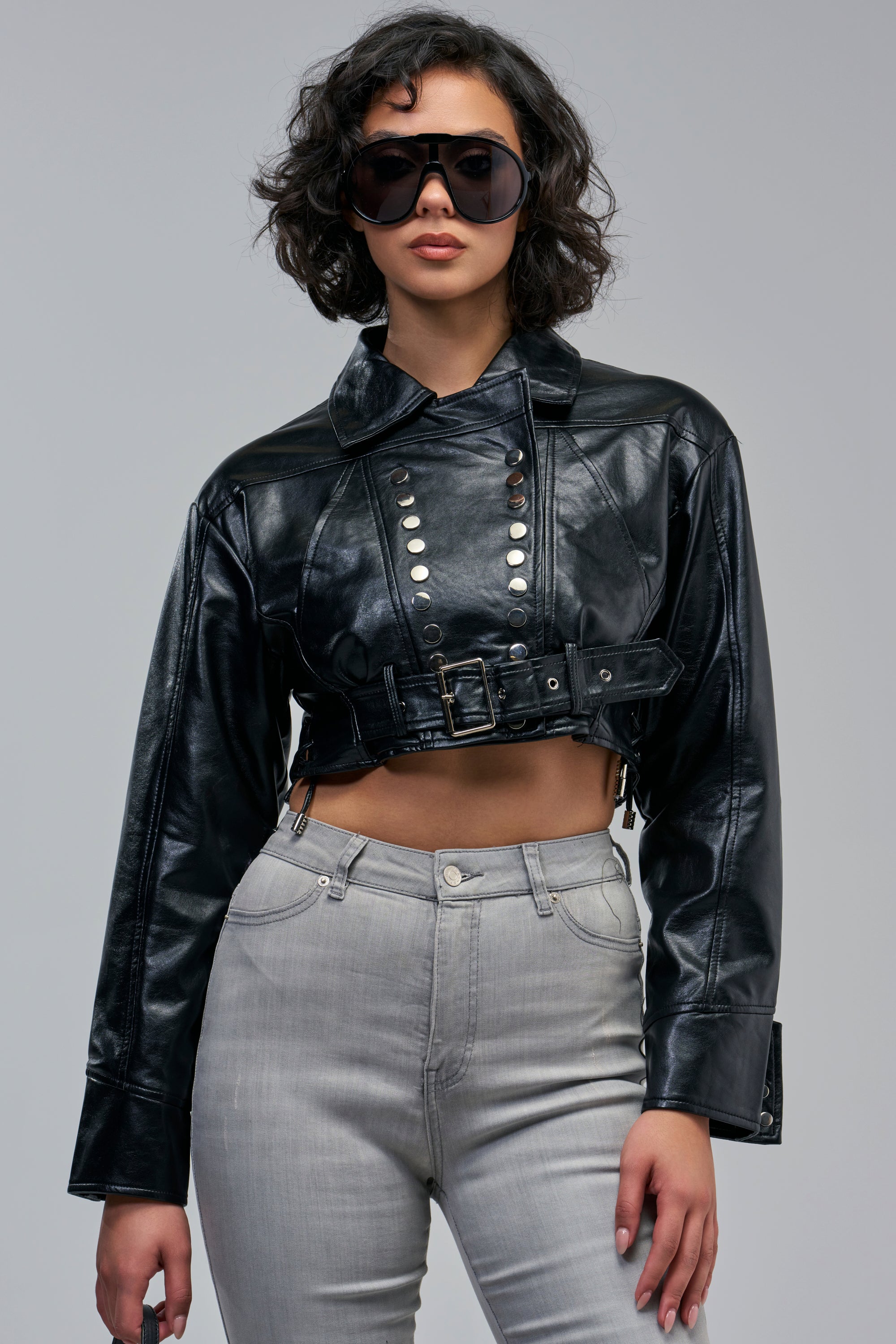 A person wearing a black faux leather JOURDAN STUDDED MOTO jacket with silver stud details and a waist belt, paired with grey jeans and dark oversized sunglasses.