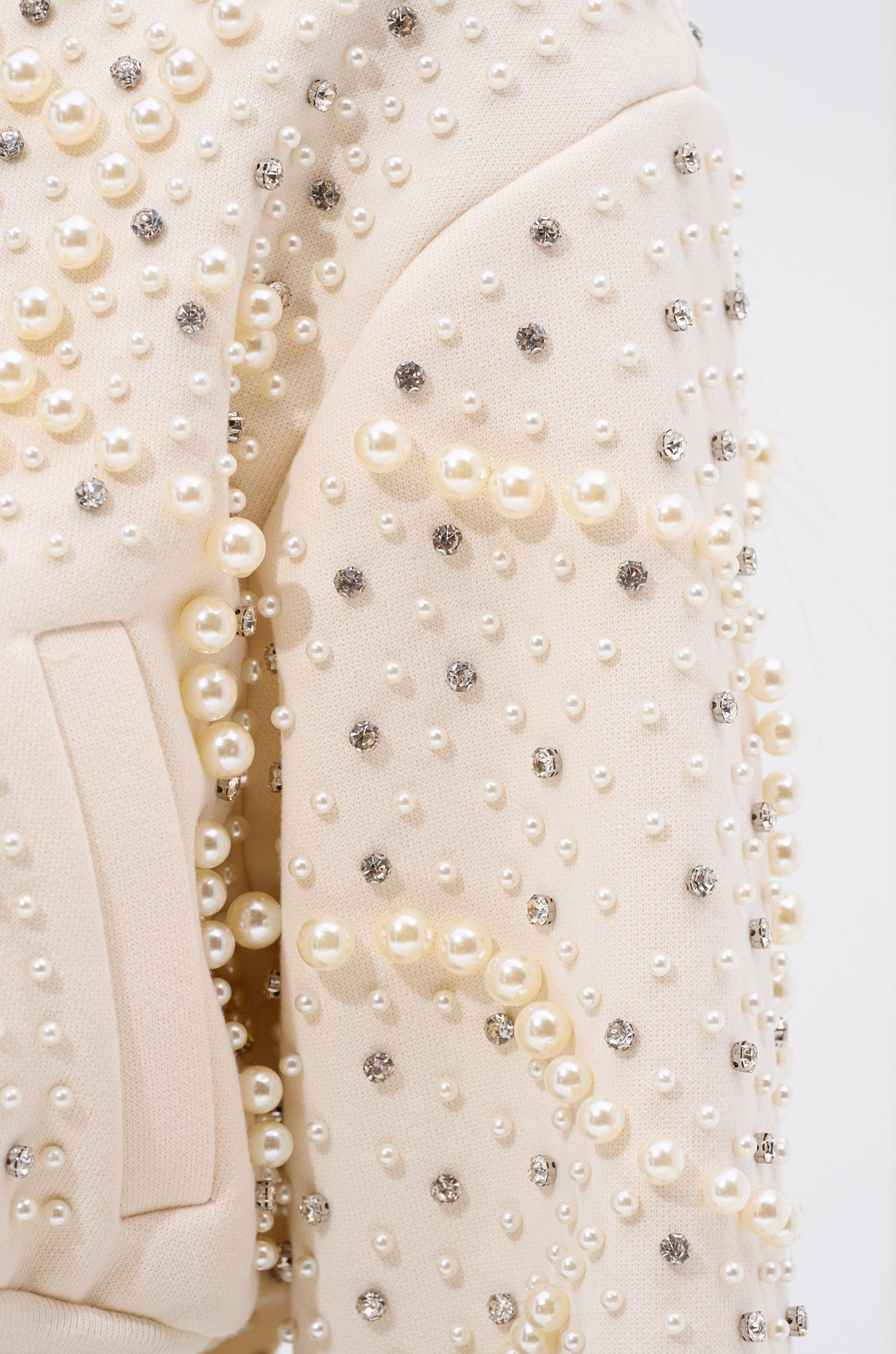 Close-up of the GOODIE TWO SHOES EMBELLISHED BOMBER sleeve, featuring clusters of pearls and rhinestones in a scattered design. A pocket seam is visible, highlighting the garment's detailed craftsmanship. 9