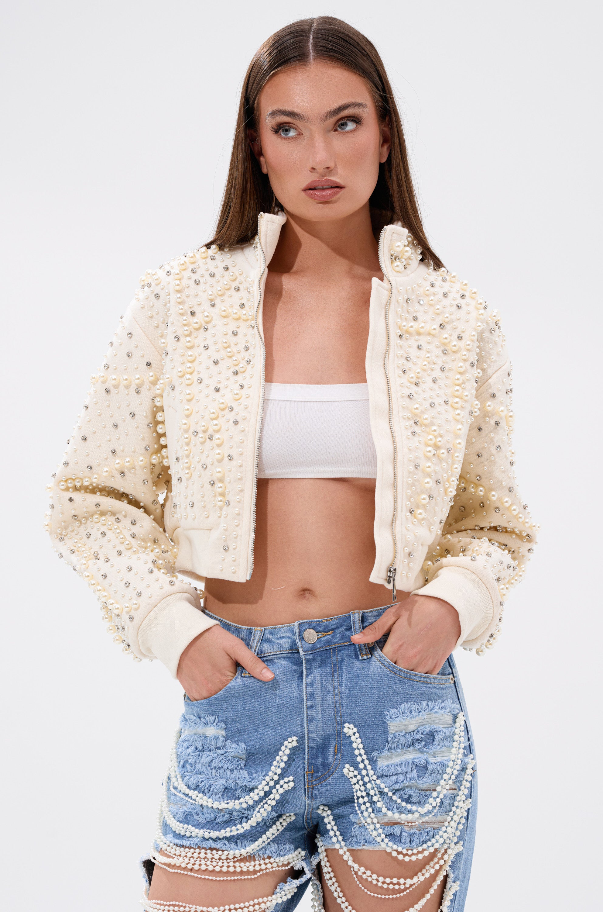 A woman stands against a plain white background wearing the GOODIE TWO SHOES EMBELLISHED BOMBER over a white crop top and high-waisted denim shorts adorned with hanging pearls. 3