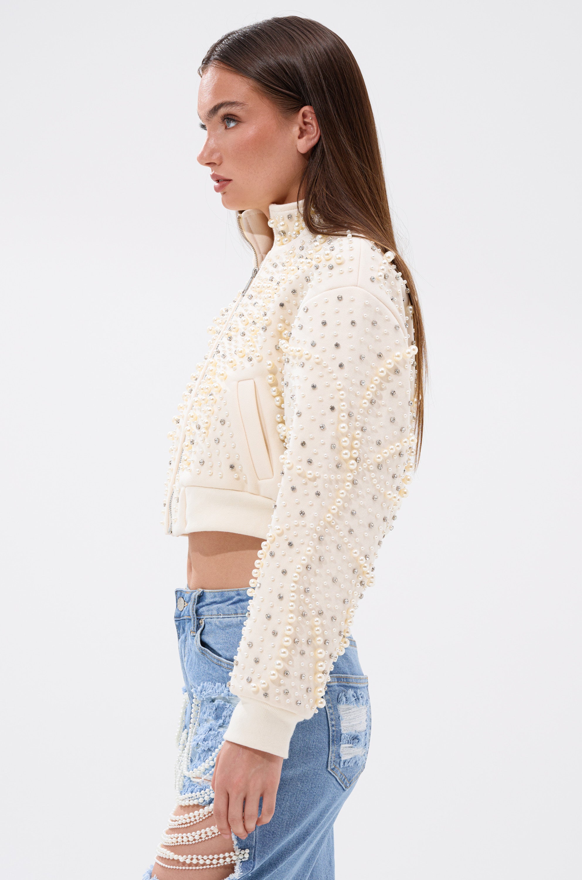 A woman in profile wears the GOODIE TWO SHOES EMBELLISHED BOMBER and light blue ripped jeans against a plain white background. 5