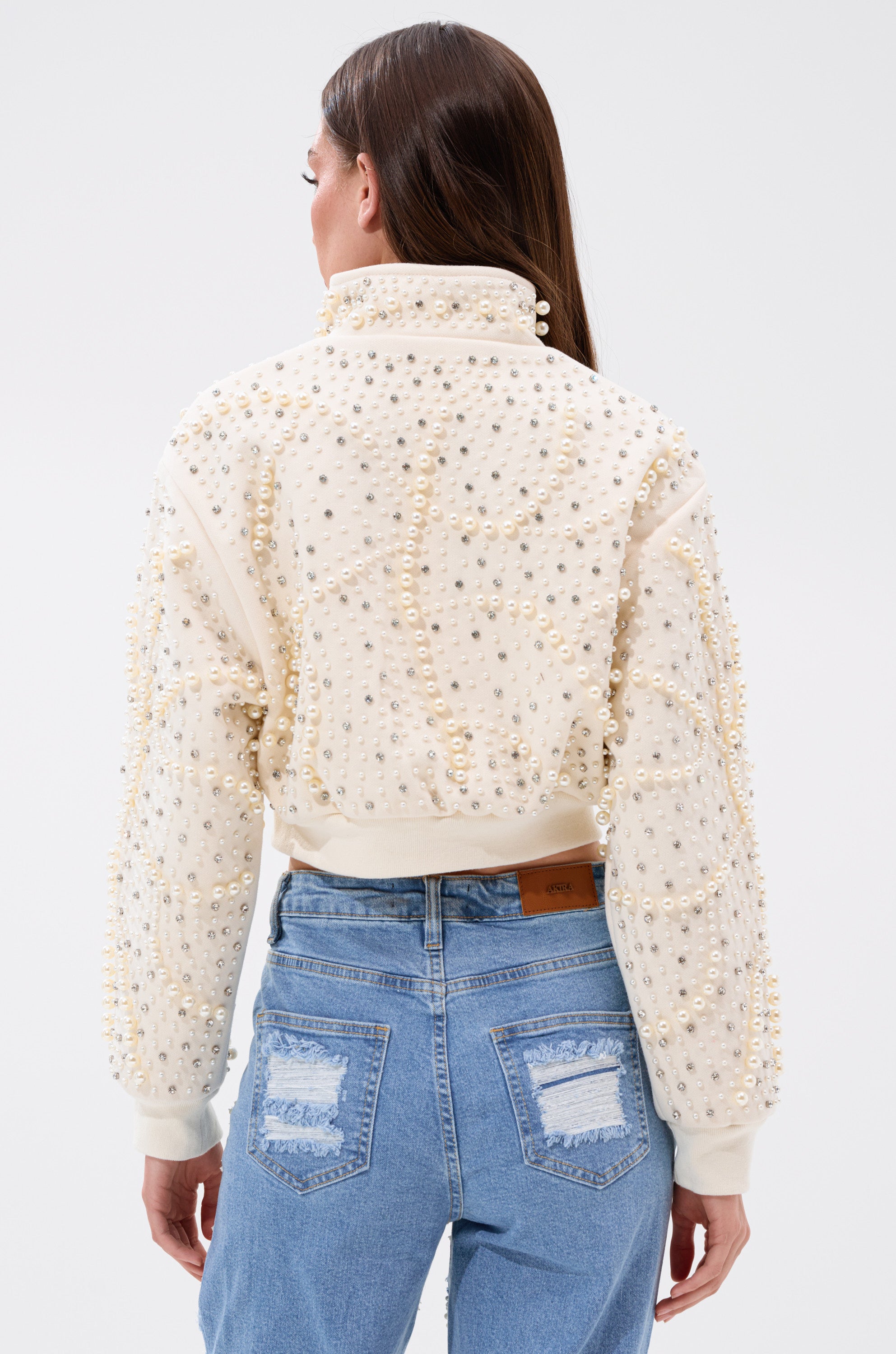 A woman with long brown hair faces away, wearing the AKIRA Label's GOODIE TWO SHOES EMBELLISHED BOMBER adorned with faux pearl rhinestone details, paired with blue high-waisted jeans featuring distressed back pockets. 6