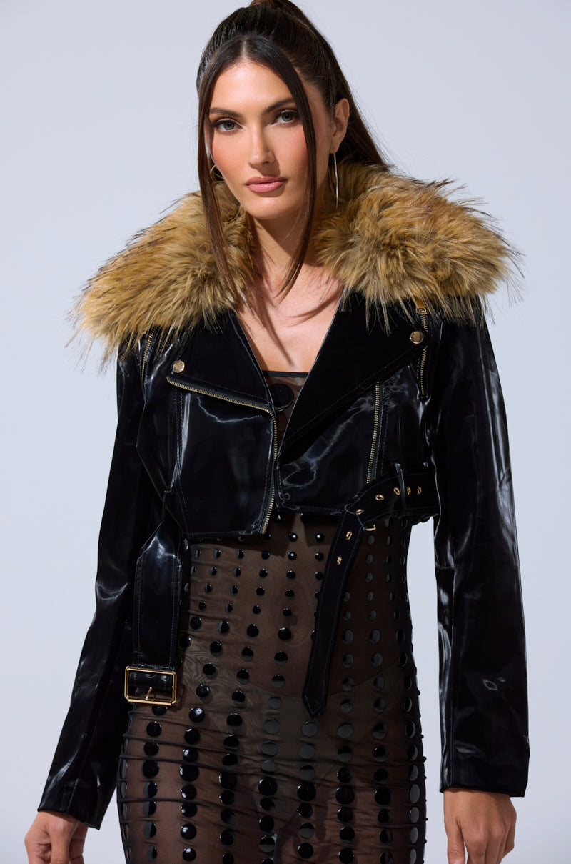 A woman with long brown hair wears the ULTRA CROP LIQUID VELVET MOTO, featuring a large removable faux fur collar, over a sheer black beaded dress. She stands against a plain light background.