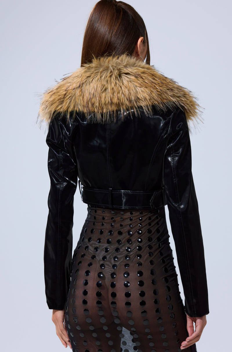 A person with long brown hair models the ULTRA CROP LIQUID VELVET MOTO jacket, featuring a removable faux fur collar, paired with a sheer black skirt with circular patterns, seen from behind against a light background.