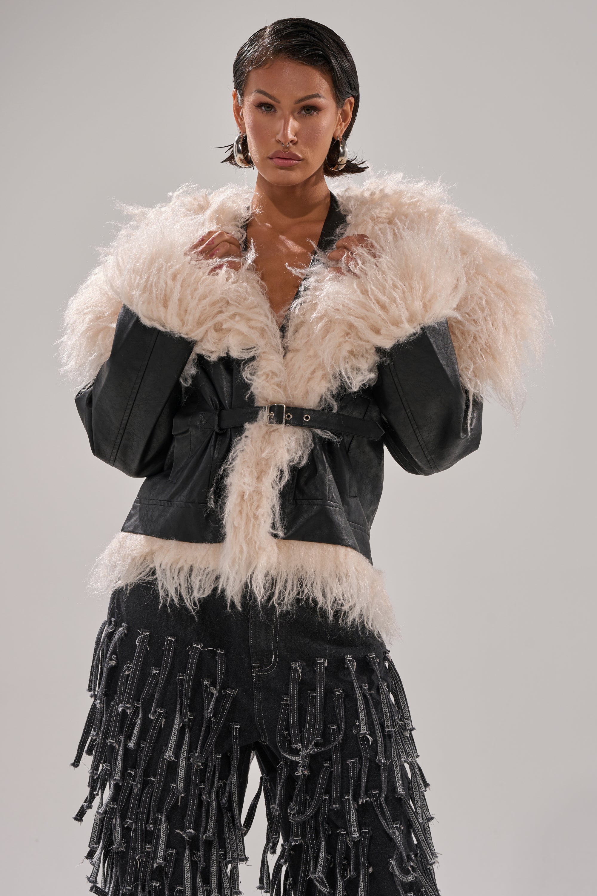 A person stands confidently against a plain gray background, wearing the ROSIE FUR TRIM MOTO—a black belted faux fur jacket with cream sherpa collar and cuffs—paired with black pants featuring multiple dangling zipper details.