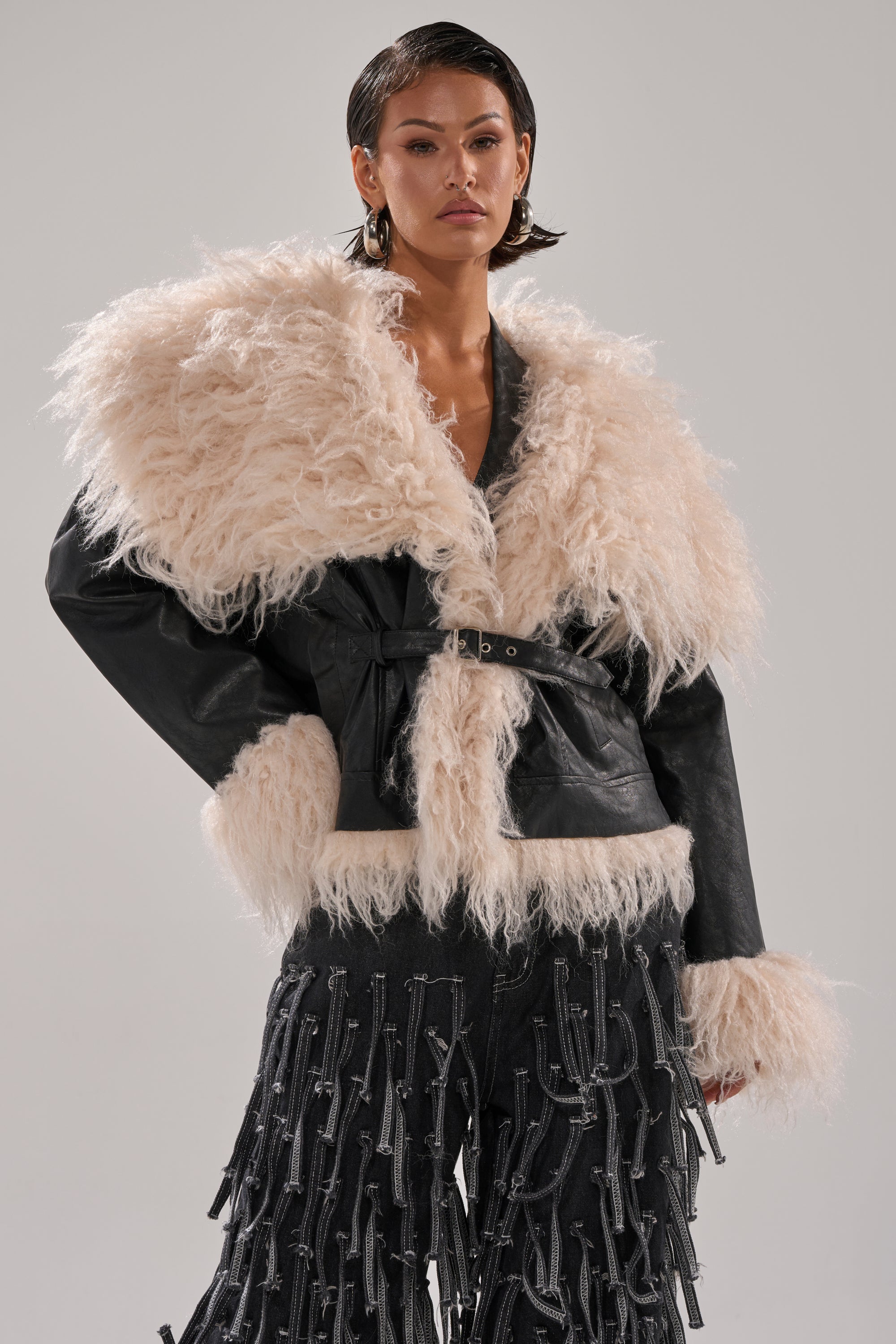 A person models the ROSIE FUR TRIM MOTO, a black faux fur moto jacket with an oversized cream shearling collar and cuffs, paired with black tassel-trimmed pants, against a plain gray background.