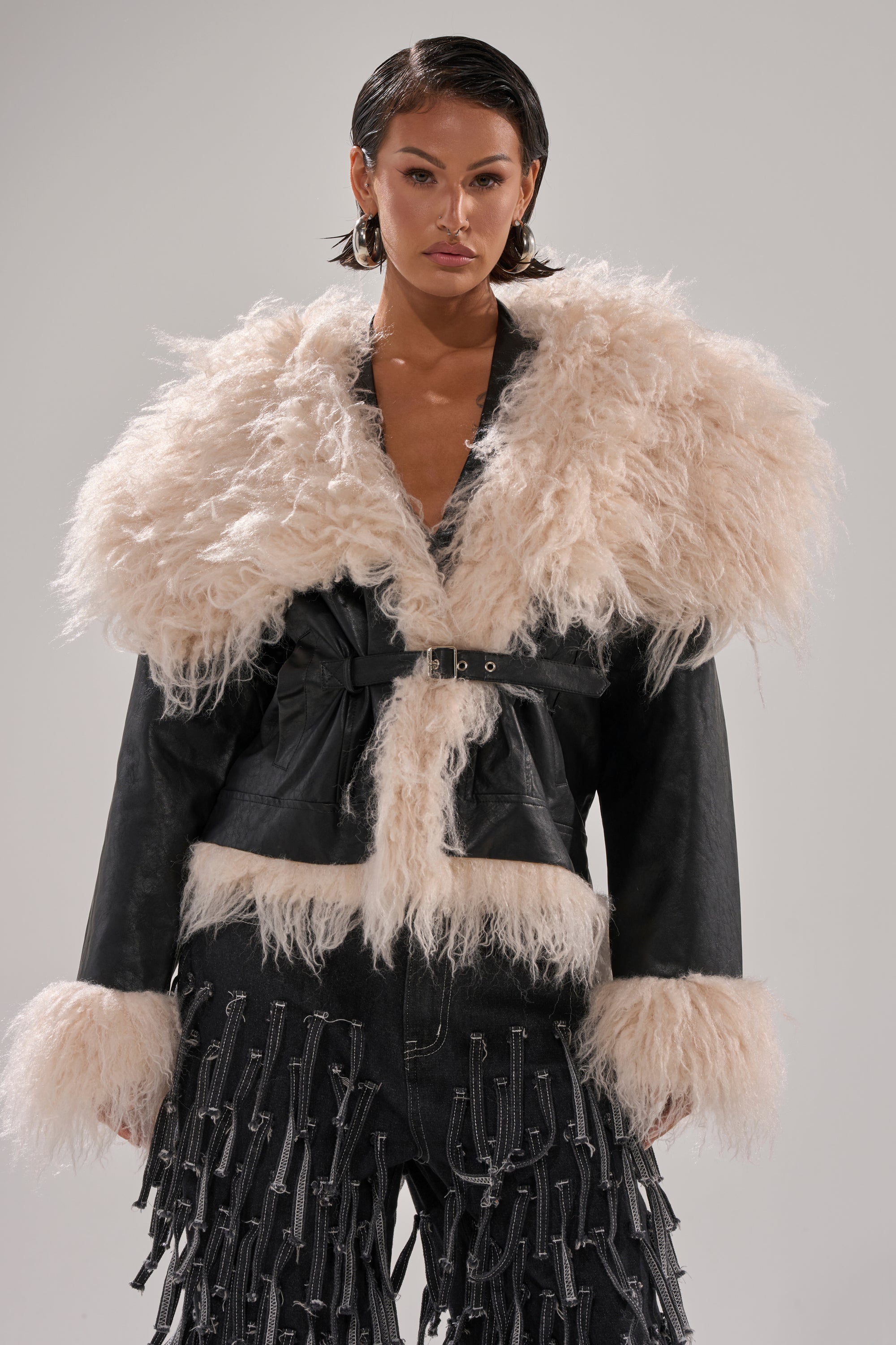 A woman models the ROSIE FUR TRIM MOTO, a black faux fur jacket with large cream-colored fur trim on the collar and cuffs, paired with black pants with chain details. She has short slicked-back hair and a neutral expression.