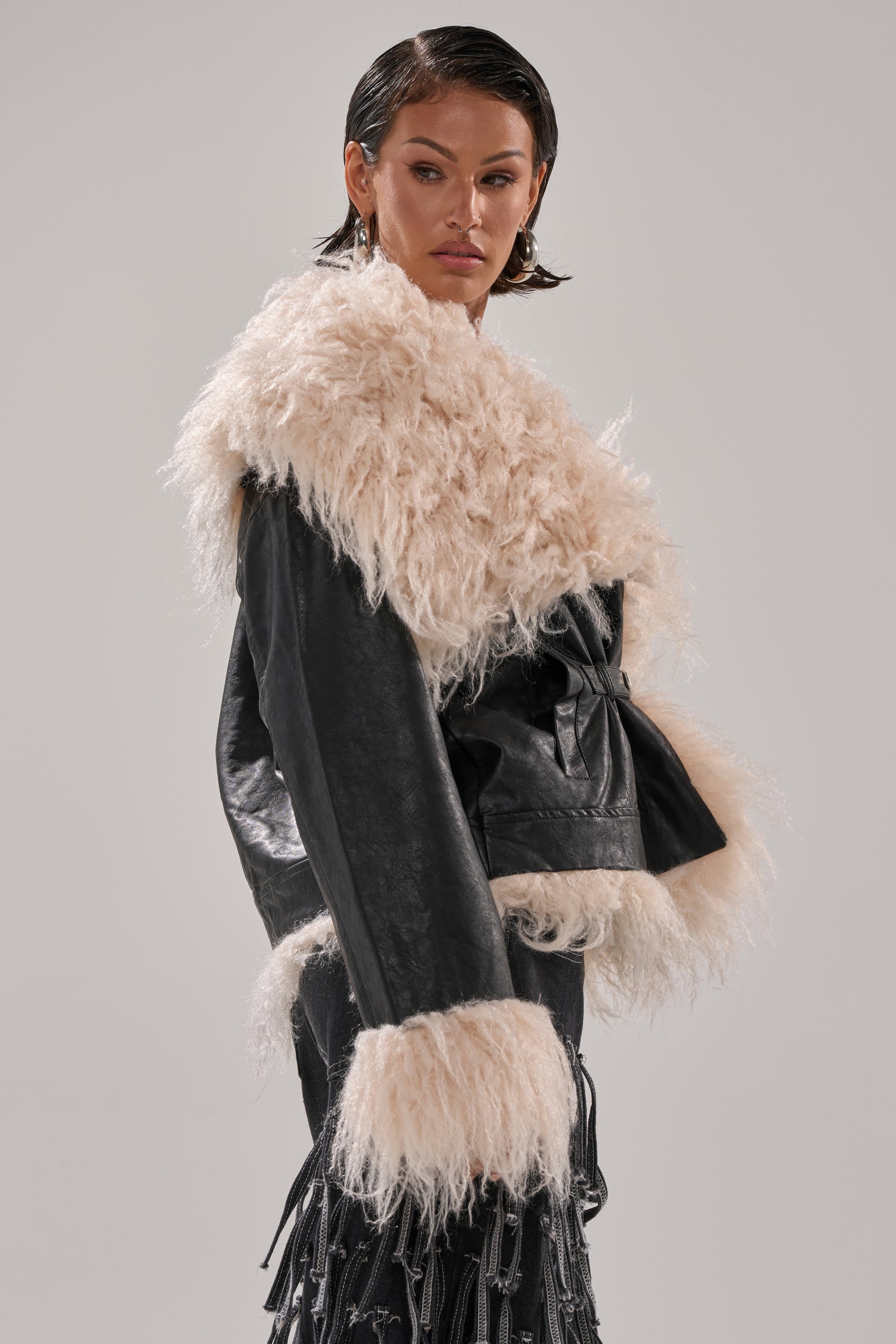 A person with slicked-back dark hair models the ROSIE FUR TRIM MOTO, a black faux fur moto jacket with bold white trim on the collar, cuffs, and edges, posing against a light gray background.