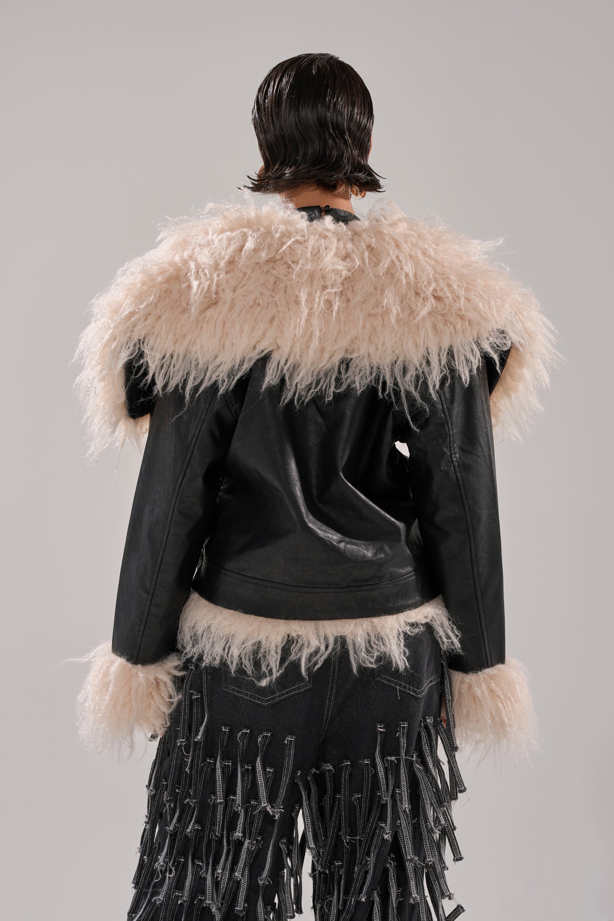 A person with short, slicked-back hair faces away, wearing the ROSIE FUR TRIM MOTO—a black faux fur jacket with fluffy beige trim on the shoulders, cuffs, and hem—paired with black pants featuring textured strips.