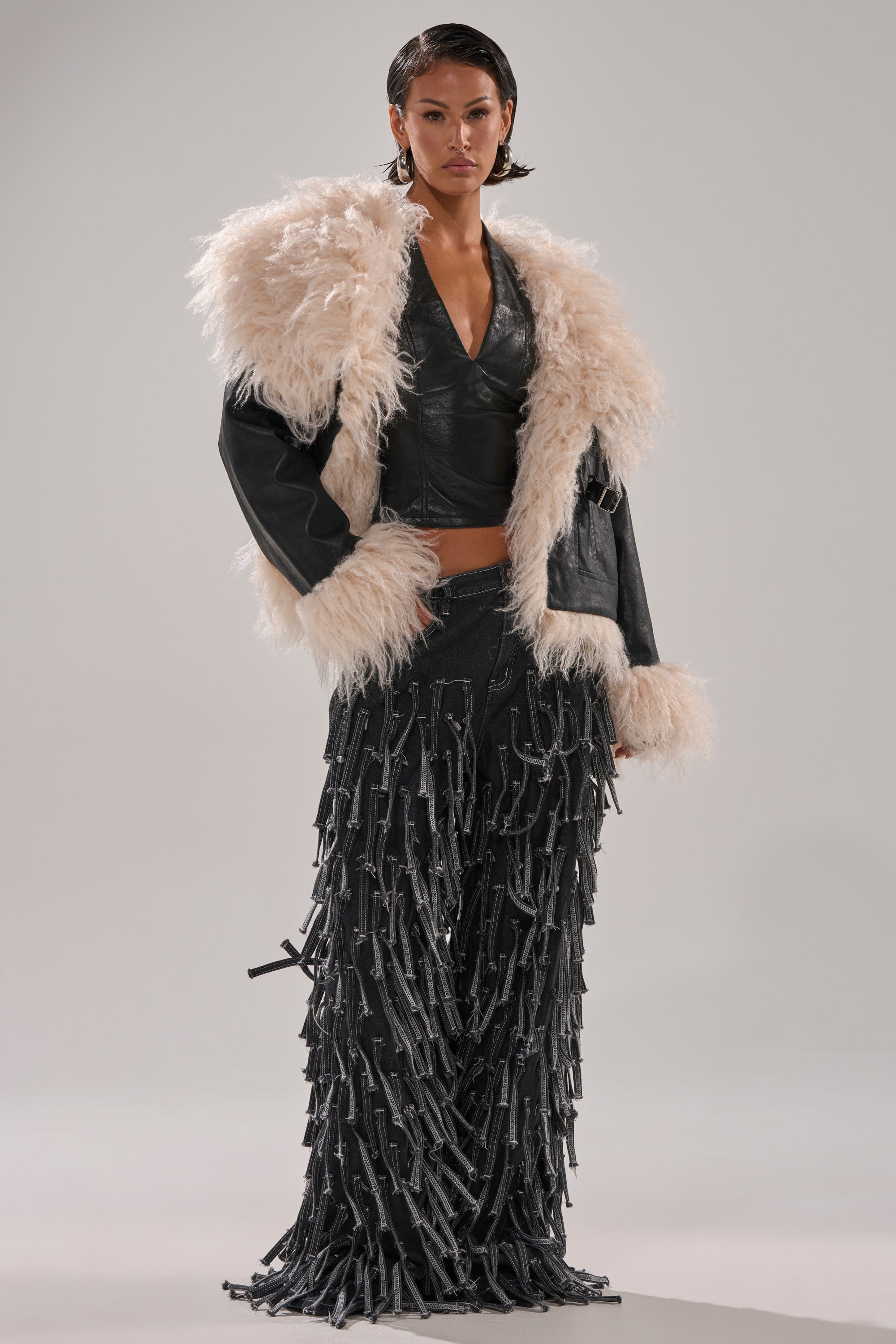 Wearing the ROSIE FUR TRIM MOTO—a faux fur moto jacket with shaggy off-white trim—over a black leather crop top and black fringe strap pants, a person stands against a plain light background.