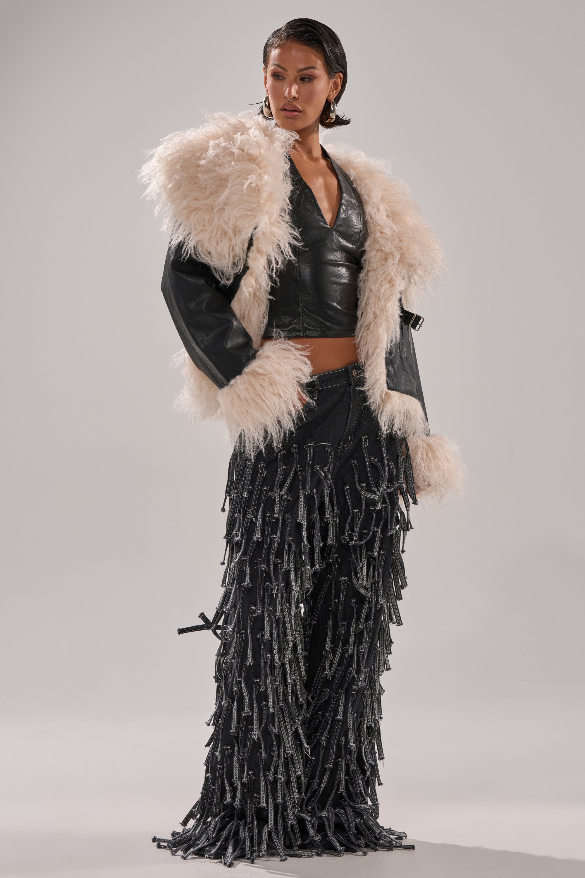 A woman poses in the ROSIE FUR TRIM MOTO jacket, featuring plush cream and black fur trim, styled with a black leather top and black pants with strap details against a plain background.