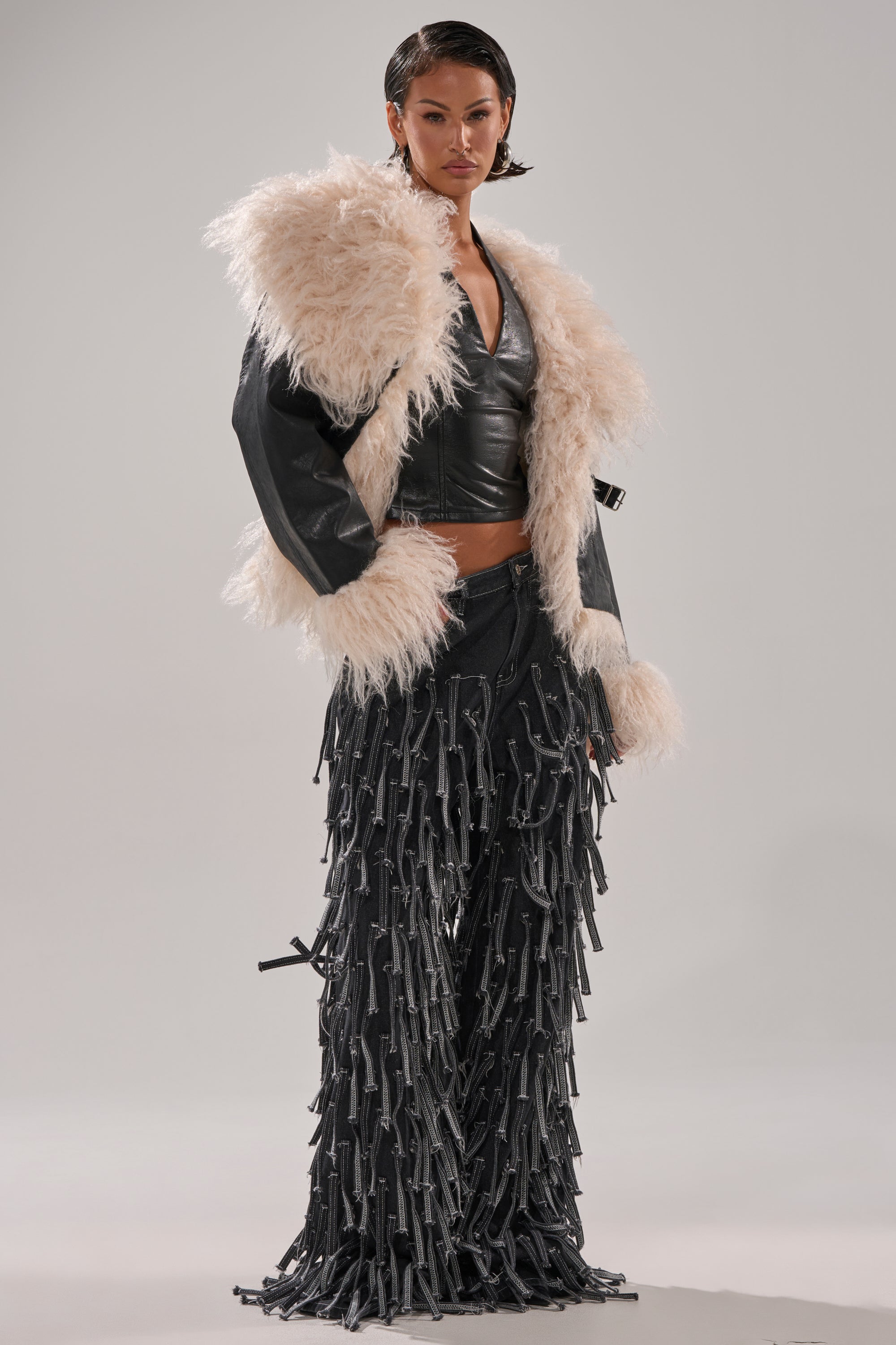 A woman poses in the ROSIE FUR TRIM MOTO, a striking faux fur moto jacket with large cream-colored trim on the collar and sleeves, styled over a black leather crop top and pants with long black fringe against a light gray background.