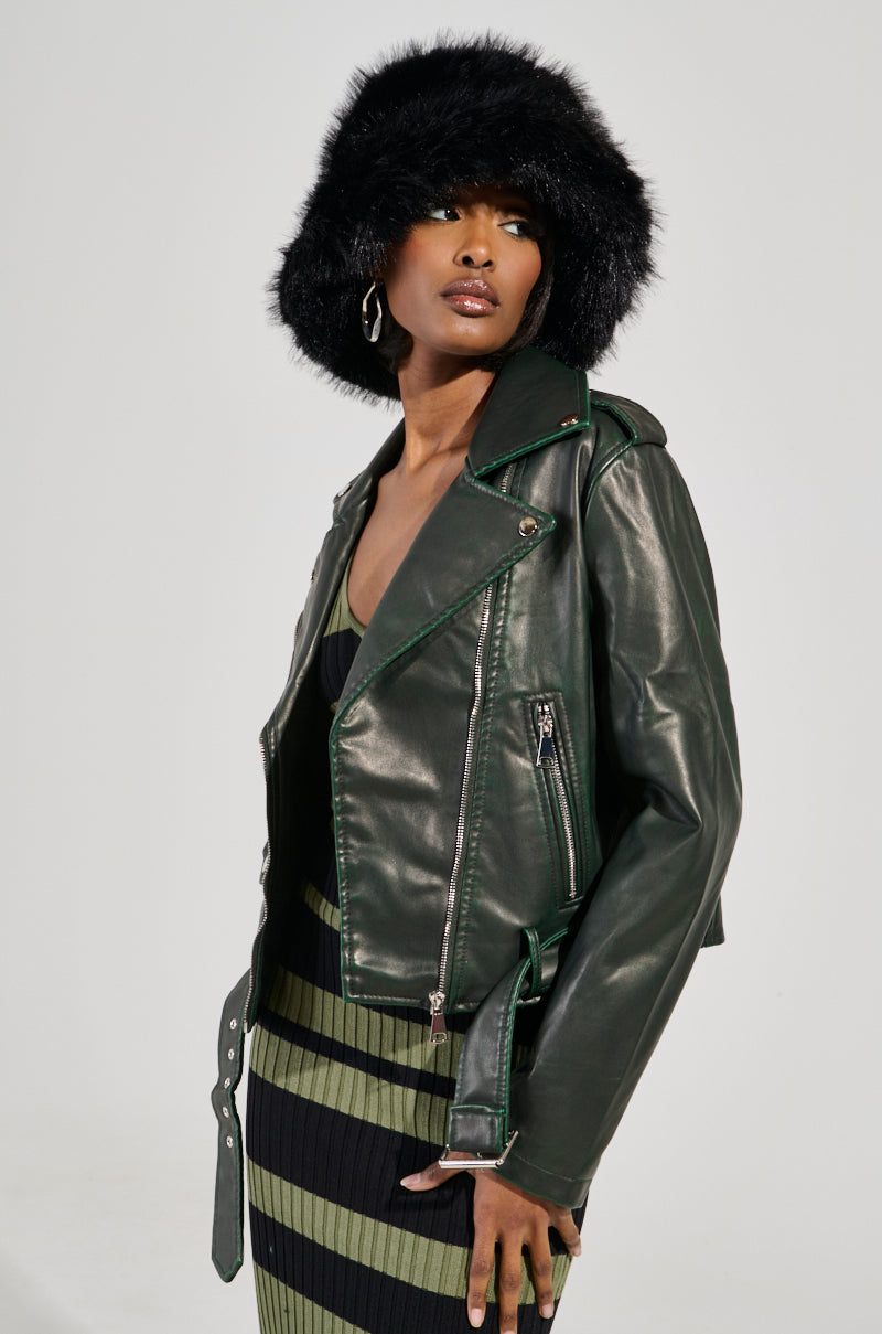 A person wearing a black fur hat, green and black striped dress, and the chic LOOKING FOR TROUBLE DISTRESSED MOTO IN GREEN jacket from the AKIRA Label poses against a light gray background. They have silver hoop earrings and are looking to the side.