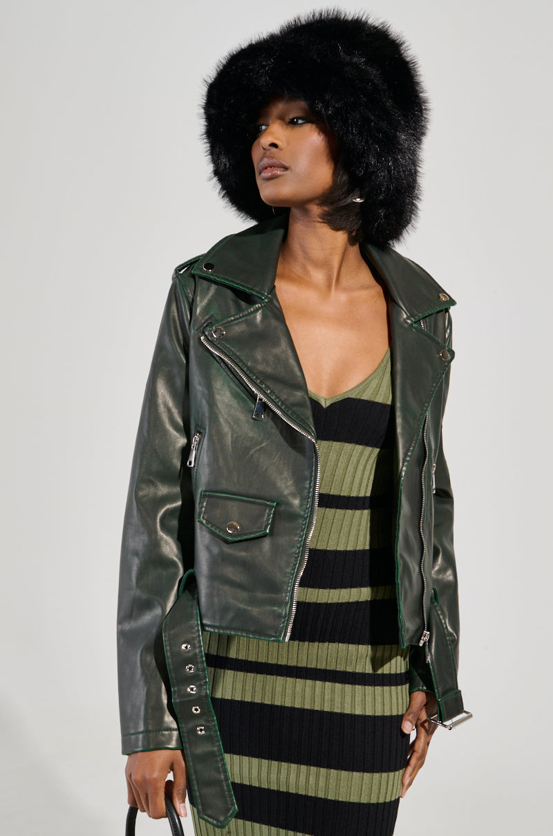 A person poses wearing an AKIRA Label black fur hat, the LOOKING FOR TROUBLE DISTRESSED MOTO IN GREEN faux leather jacket, and a green and black striped dress. They hold a handbag and stand against a neutral background.