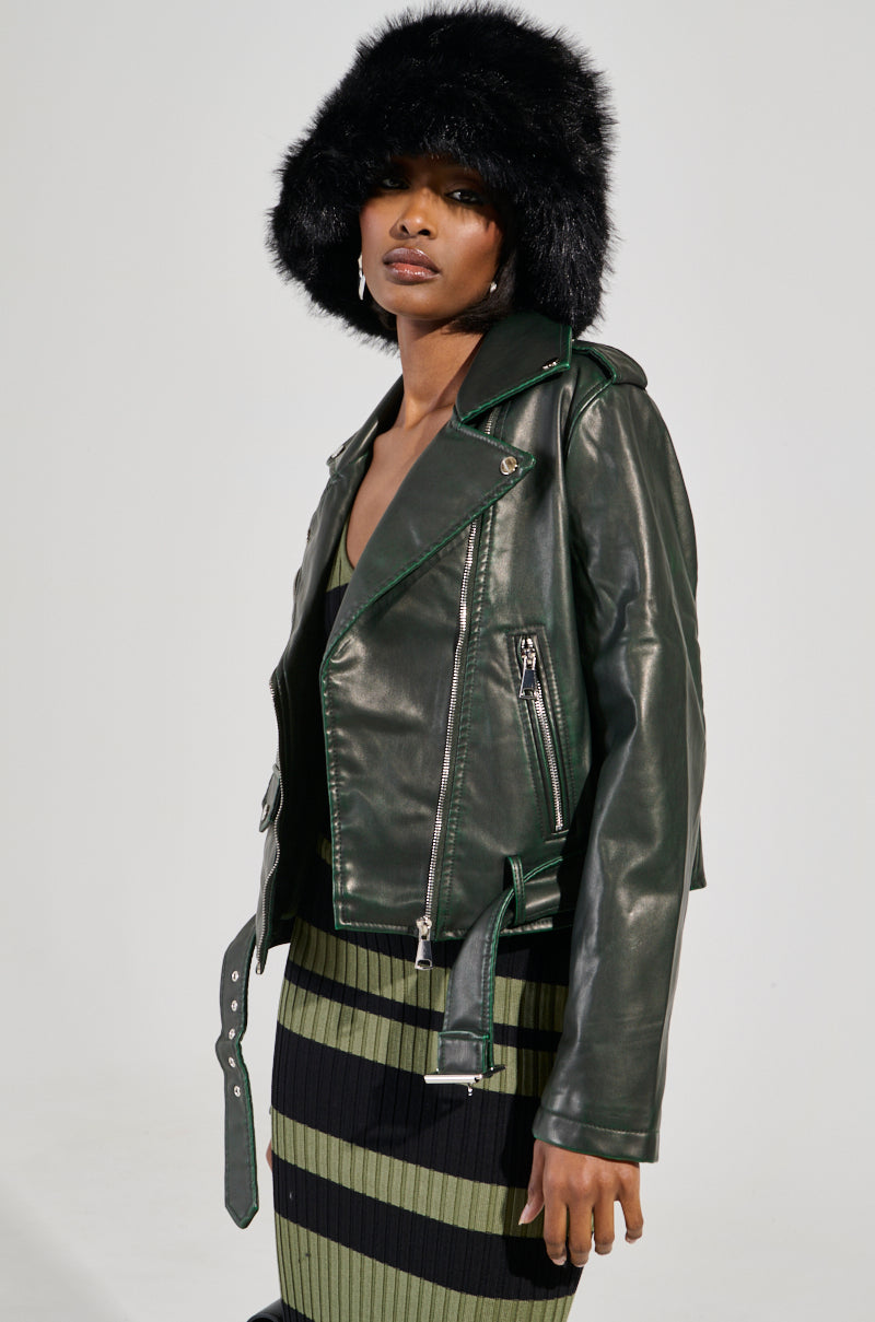 A person is dressed in the 'Looking For Trouble Distressed Moto' jacket in green, accompanied by a furry black hat and a green-and-black striped skirt. The light, plain background emphasizes their fashionable ensemble.