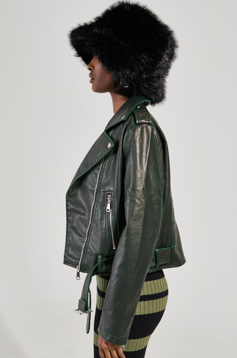 A person stands in profile wearing a black faux fur hat, an AKIRA Label faux leather jacket, and the LOOKING FOR TROUBLE DISTRESSED MOTO IN GREEN skirt. The outfit exudes a modern and stylish appearance against a plain, light-colored background.