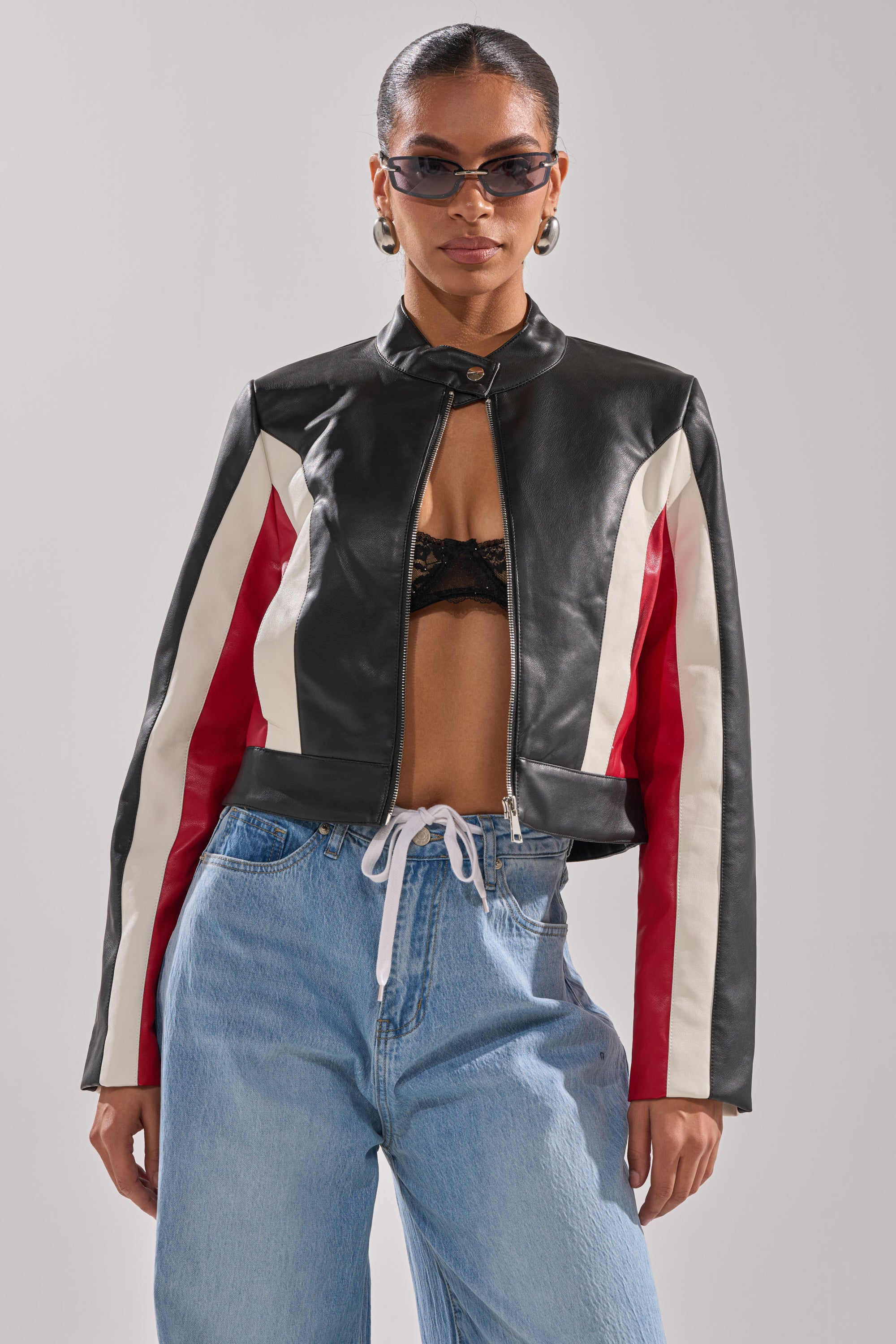 A woman stands confidently in the ON YOUR MARKS RACER MOTO jacket—faux leather with red and white stripes—styled with sunglasses, a black lace bralette, and light blue jeans featuring a white drawstring against a plain background. 0
