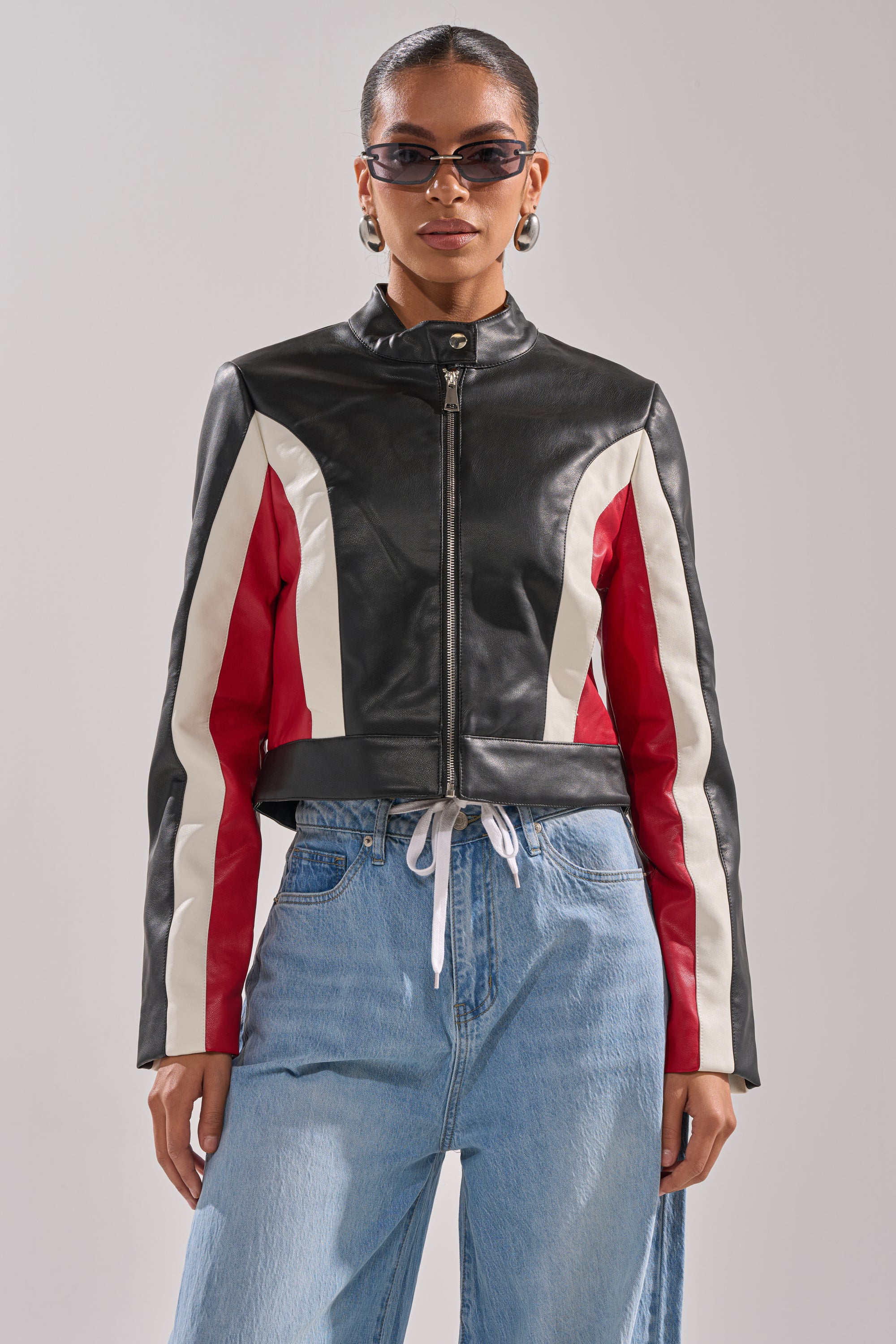 A woman wearing sunglasses, gold hoop earrings, and the ON YOUR MARKS RACER MOTO black color block jacket over a white top and loose blue jeans stands facing forward against a plain light background. 1