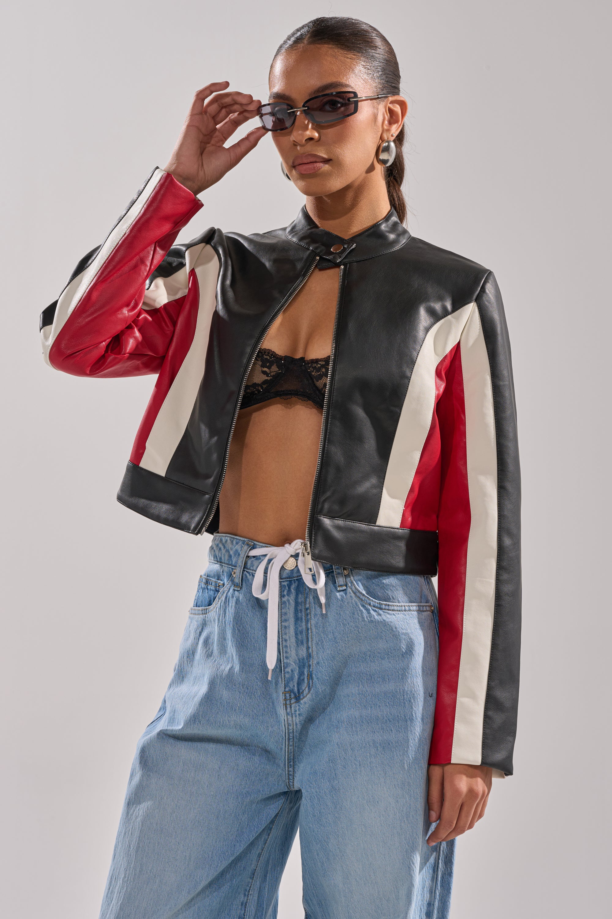 A woman stands confidently against a plain background wearing the ON YOUR MARKS RACER MOTO jacket over a black lace bralette, paired with light blue drawstring jeans and sunglasses. 2
