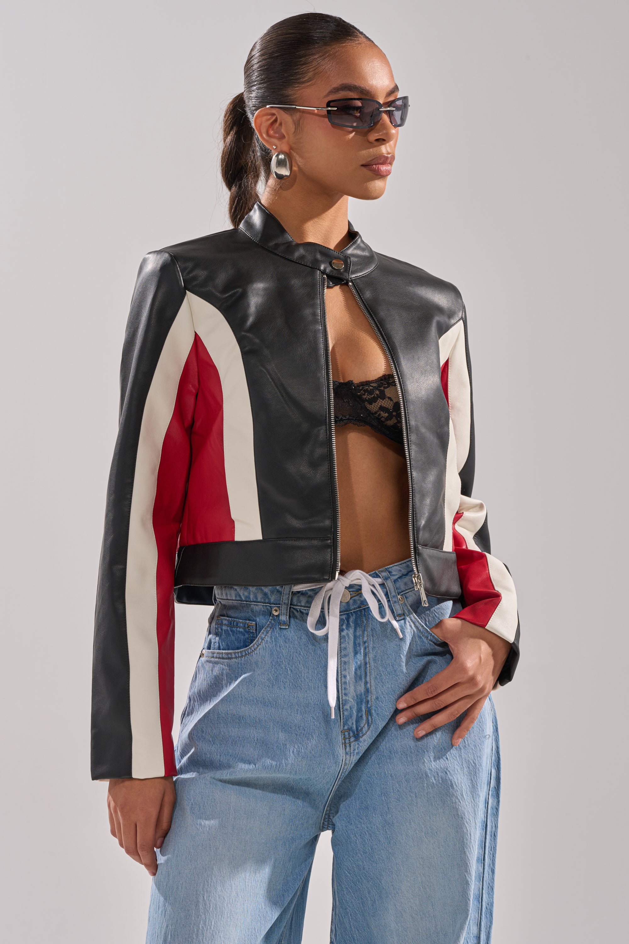 A woman poses confidently against a plain background wearing sunglasses, hoop earrings, and the ON YOUR MARKS RACER MOTO black color block jacket with white and red stripes, styled with a black lace bralette and light blue jeans with a white drawstring. 3