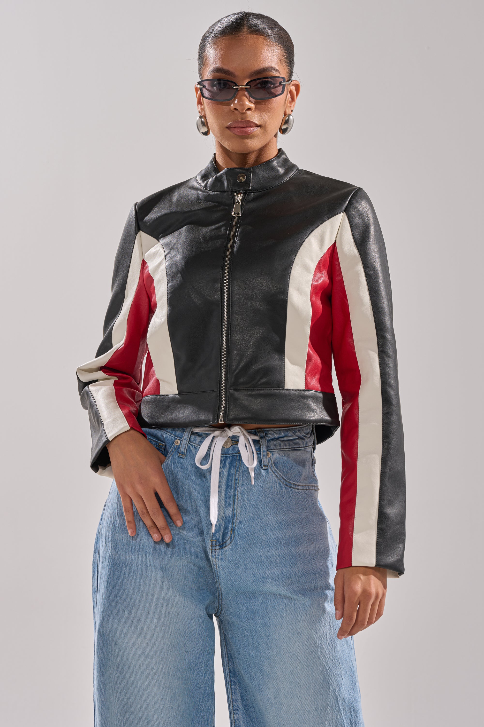 A woman models the ON YOUR MARKS RACER MOTO jacket—featuring white and red stripes—paired with sunglasses and light blue jeans, posing confidently against a plain background. 4