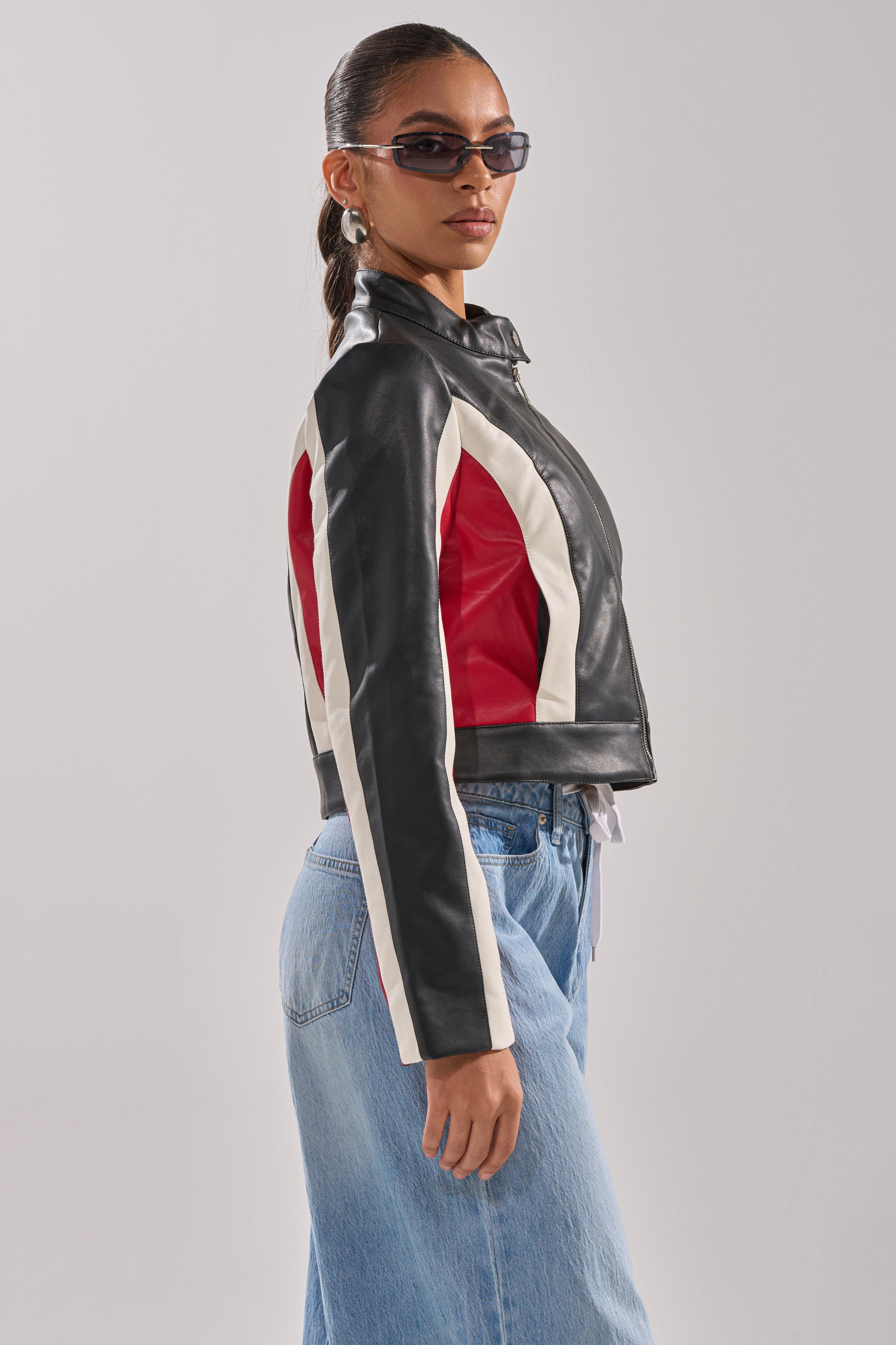 A woman wearing sunglasses and the ON YOUR MARKS RACER MOTO jacket with white and red stripes, paired with light blue jeans, stands sideways against a plain light background. 5