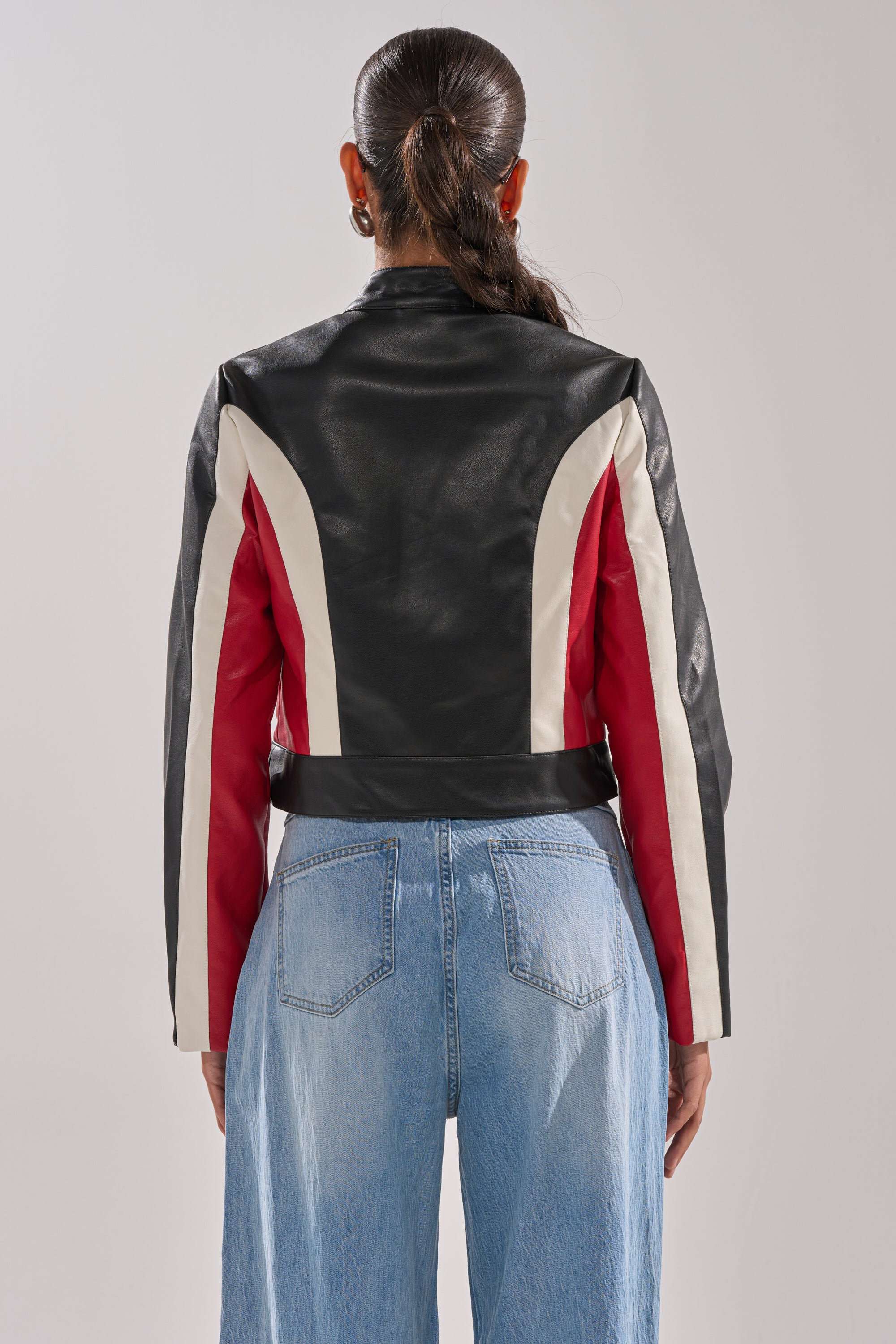 A woman with long dark hair tied back is seen from behind wearing the ON YOUR MARKS RACER MOTO women's color block jacket in black, white, and red with blue jeans against a plain light background. 6