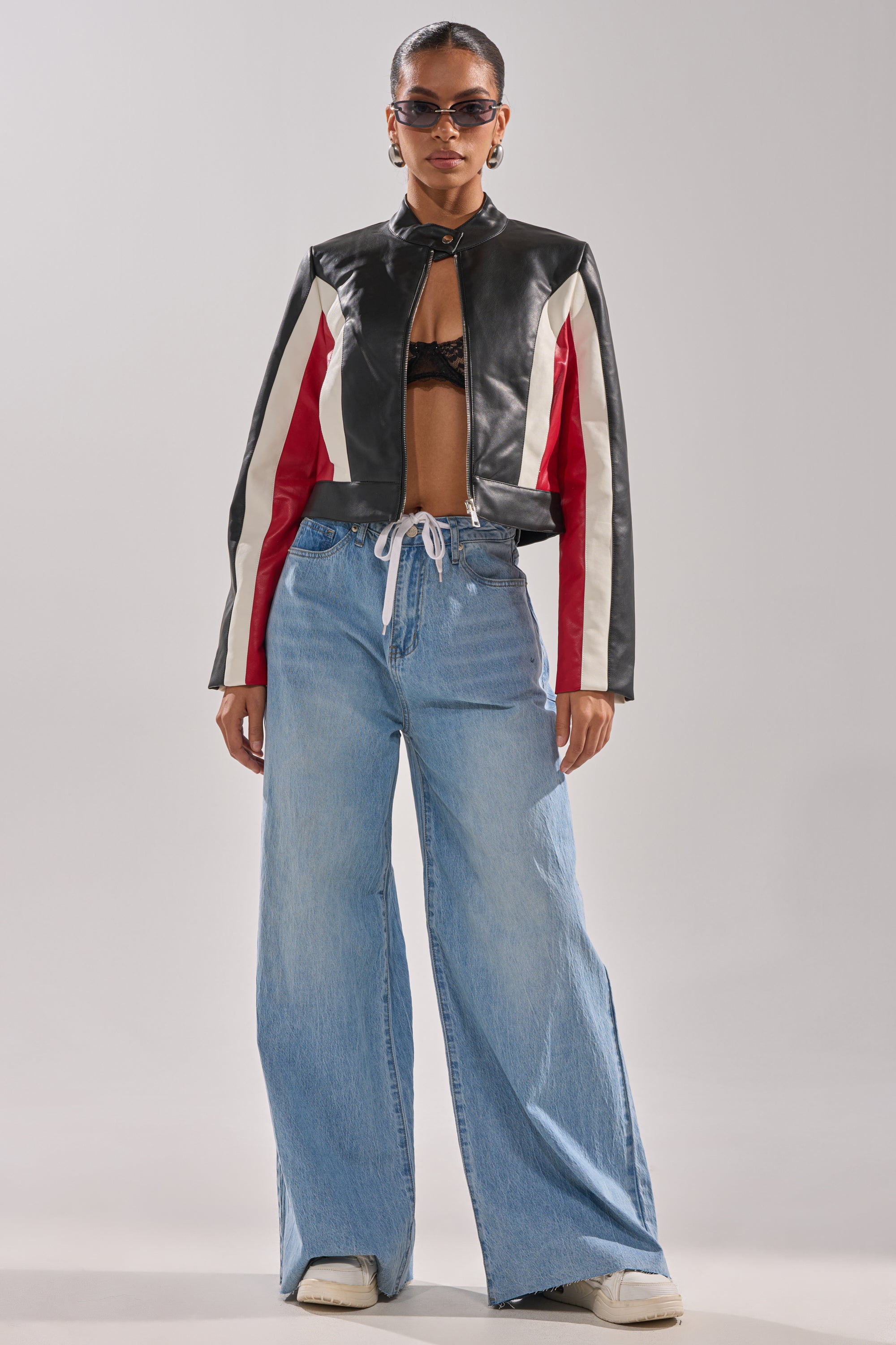 A person wearing sunglasses, the ON YOUR MARKS RACER MOTO cropped black faux leather jacket with white and red stripes, a black bra top, wide-leg light blue drawstring jeans, and white sneakers stands against a plain background. 7