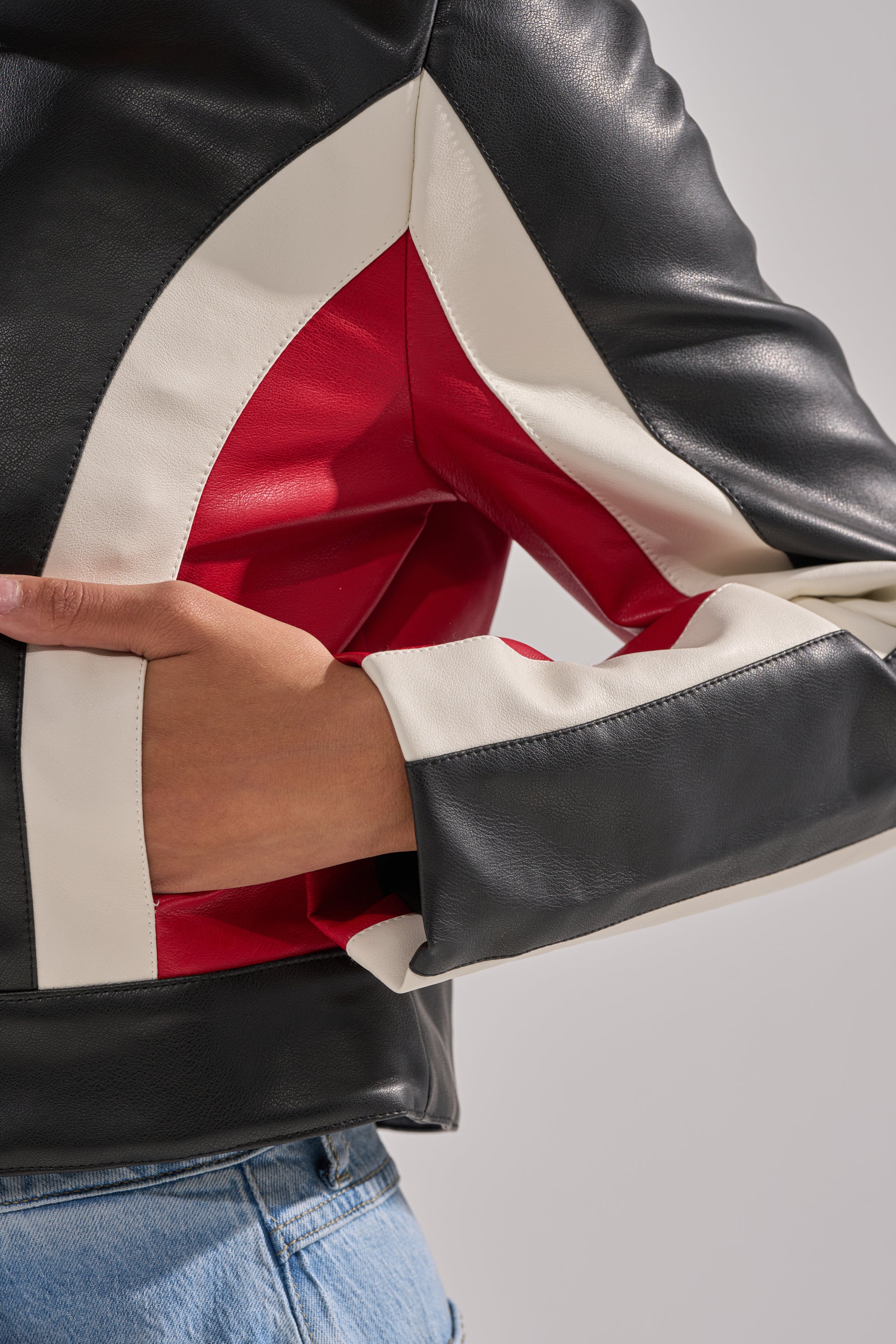 A close-up of a person wearing the ON YOUR MARKS RACER MOTO jacket—black, white, and red geometric panels in faux leather—paired with blue jeans, with their hand resting in the pocket. 8