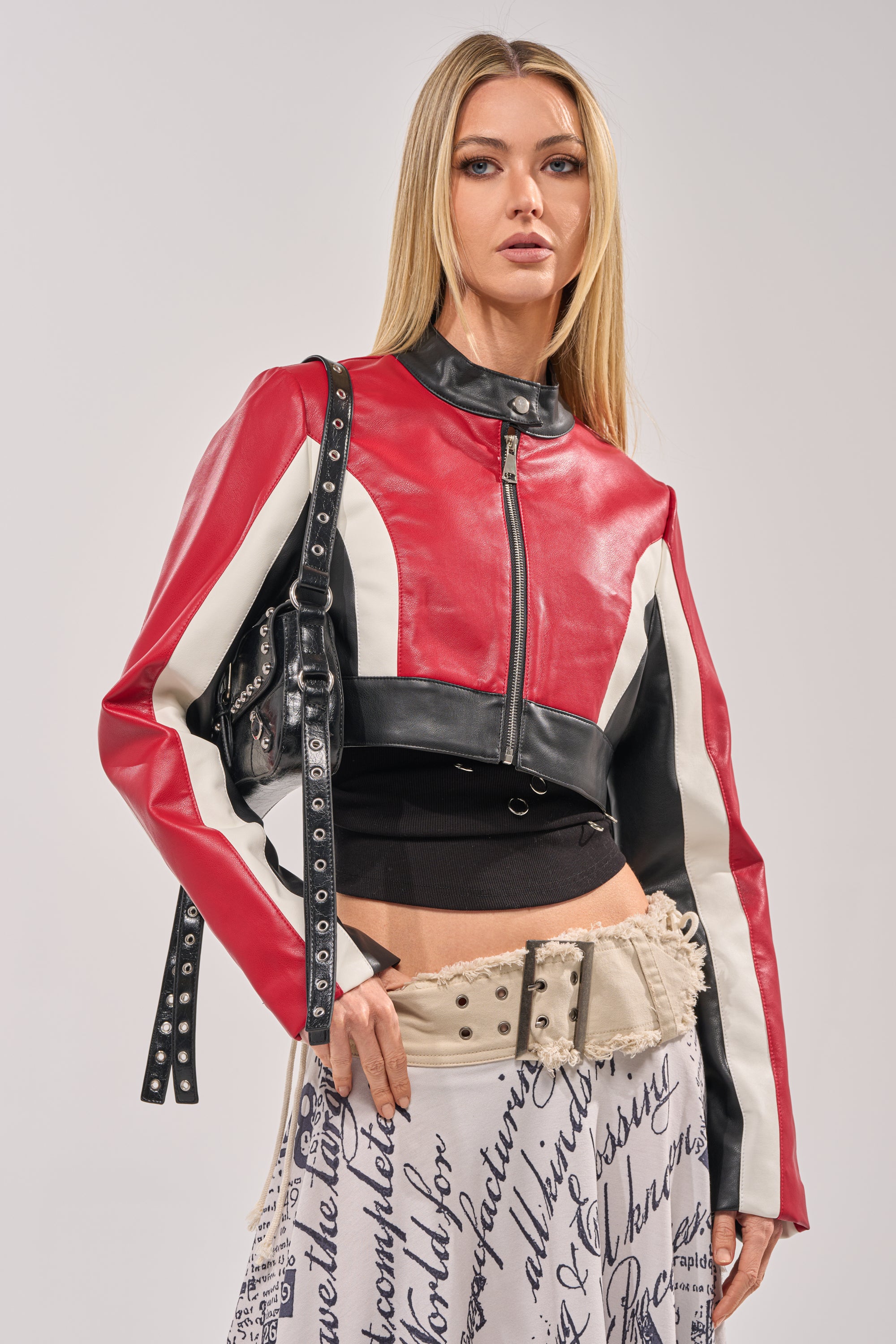 A woman with long blonde hair wears the ON YOUR MARKS CROPPED MOTO jacket, a studded black bag, black top, and a cream skirt with black cursive writing as she poses against a plain light background. 0