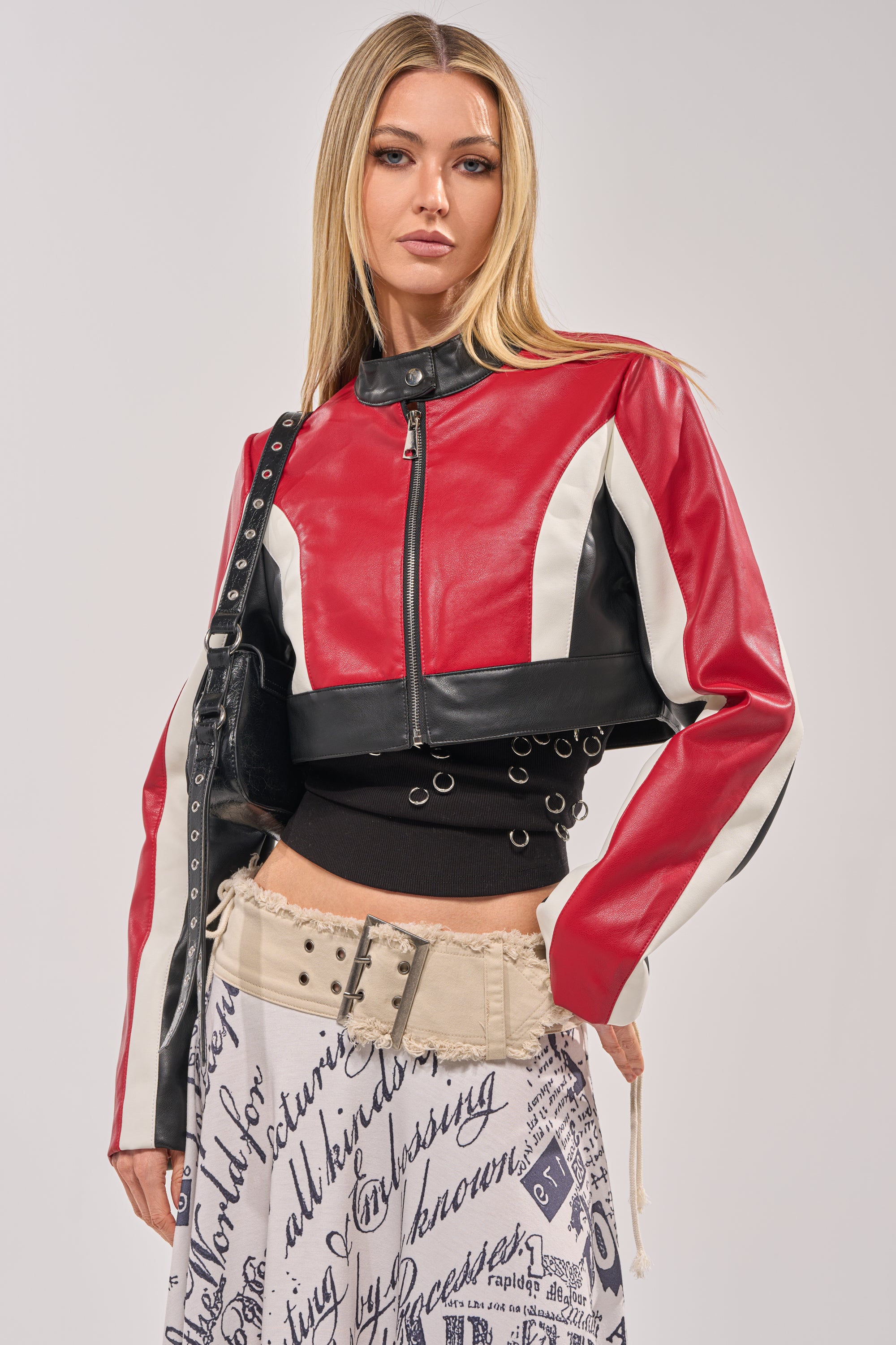 A woman with straight blonde hair wears the ON YOUR MARKS CROPPED MOTO red faux leather jacket, a black grommet crop top, and a beige graphic skirt, paired with a black studded bag—an ideal girls' night out look. 3