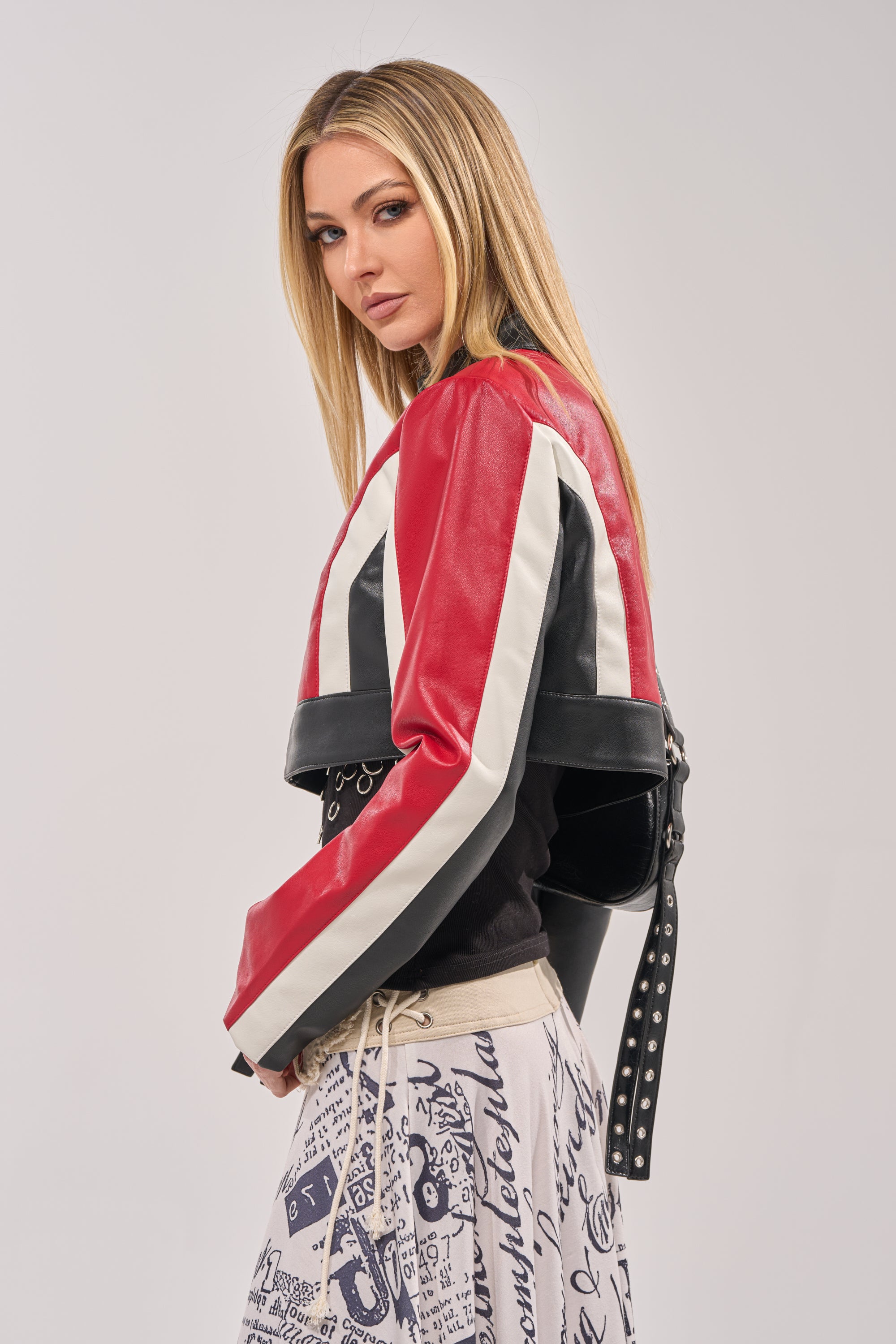 A woman with long blonde hair wears the ON YOUR MARKS CROPPED MOTO, a red colorblock faux leather jacket, over a black top and a white skirt with black graphic designs. She poses sideways, gazing confidently at the camera. 4