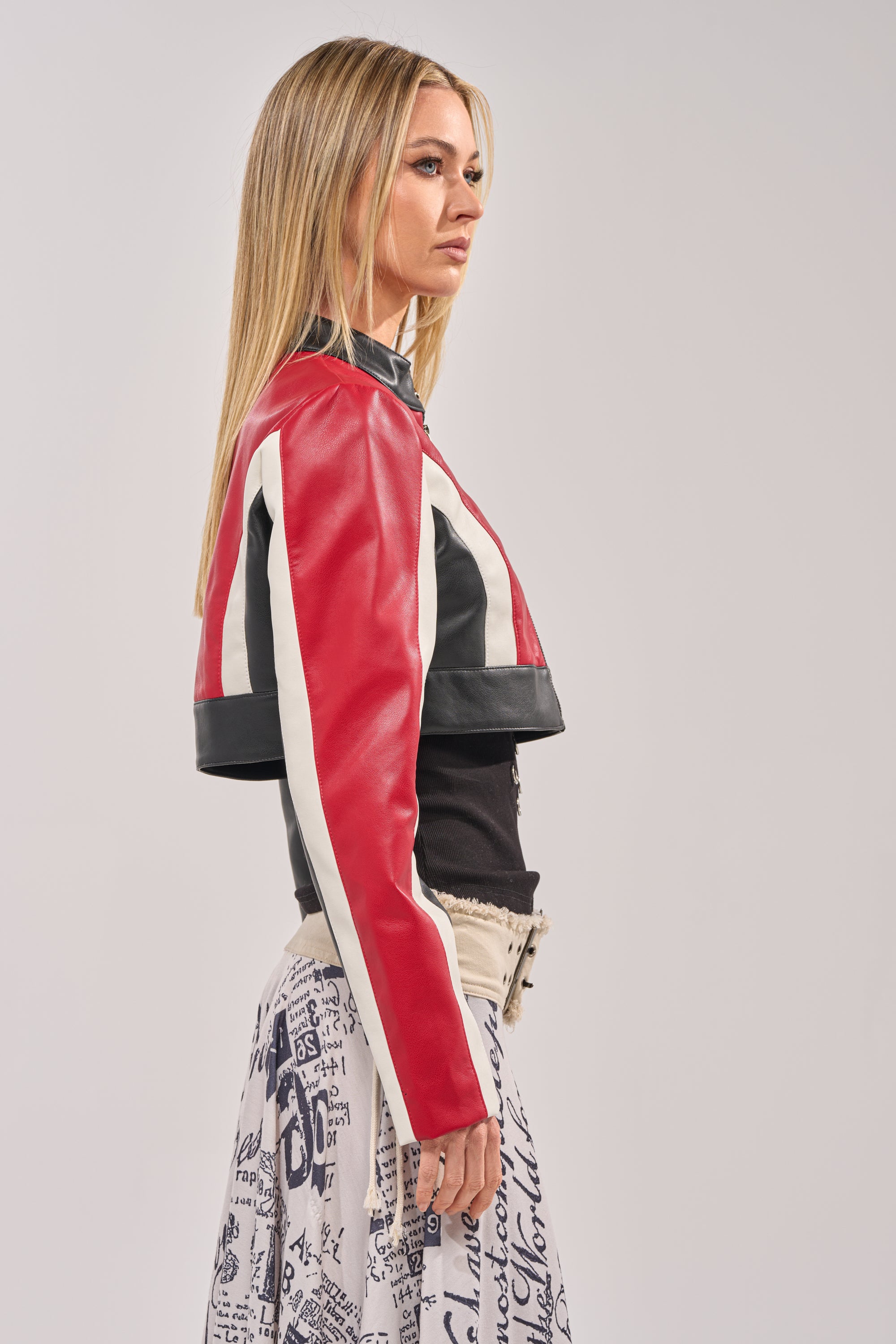 A woman with long blonde hair stands in profile, wearing the ON YOUR MARKS CROPPED MOTO jacket in red over a black top and a white skirt with black graphic text, against a plain light background. 5
