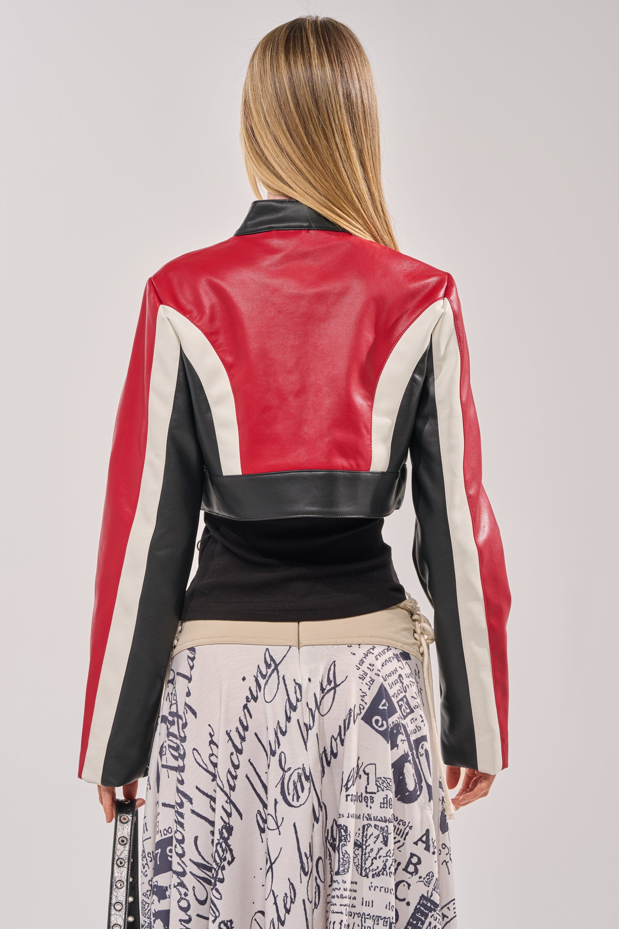 A woman with long blonde hair, seen from behind, wears the ON YOUR MARKS CROPPED MOTO—a red faux leather colorblock jacket—paired with a skirt featuring black handwritten-style text and graphics on a light background. 6