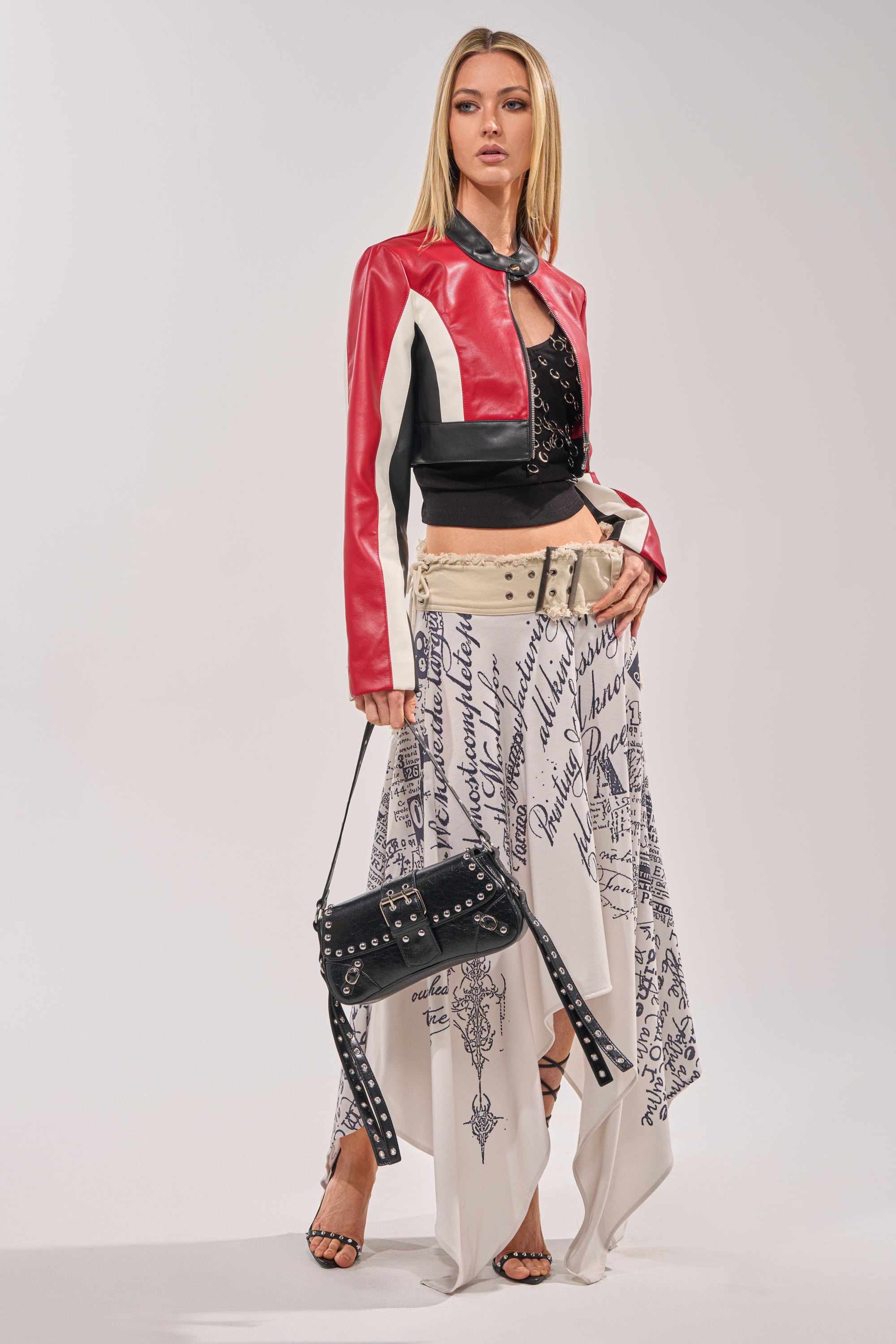 A woman stands confidently in the ON YOUR MARKS CROPPED MOTO— a red and black colorblock jacket—paired with a black top, white script-print skirt, strappy heels, and a studded black handbag against a plain background. 7