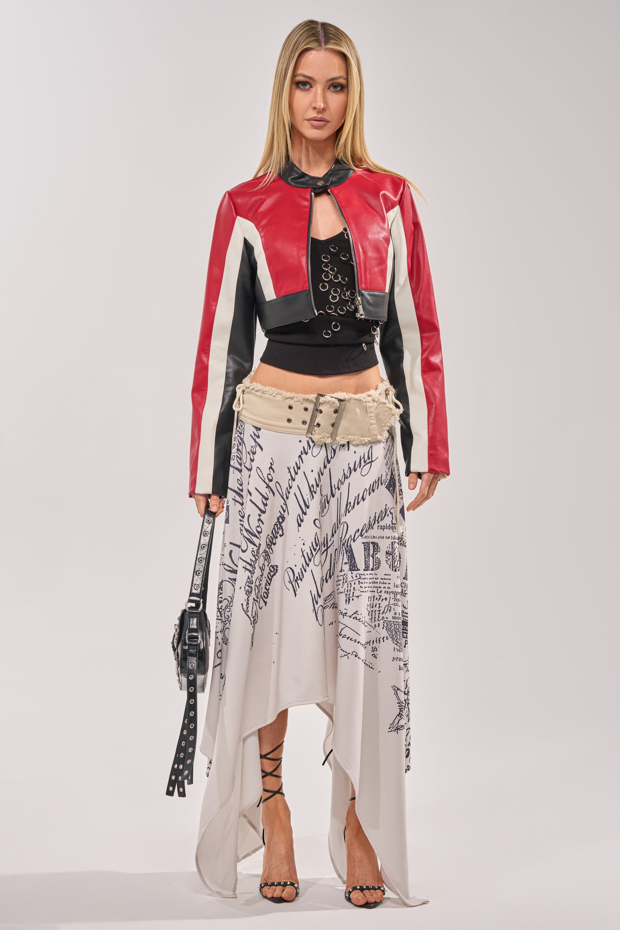 A woman faces forward in the ON YOUR MARKS CROPPED MOTO jacket over a black top, paired with a white asymmetric skirt featuring black script, strappy heels, and a black studded bag—ideal for a girls' night out. 8