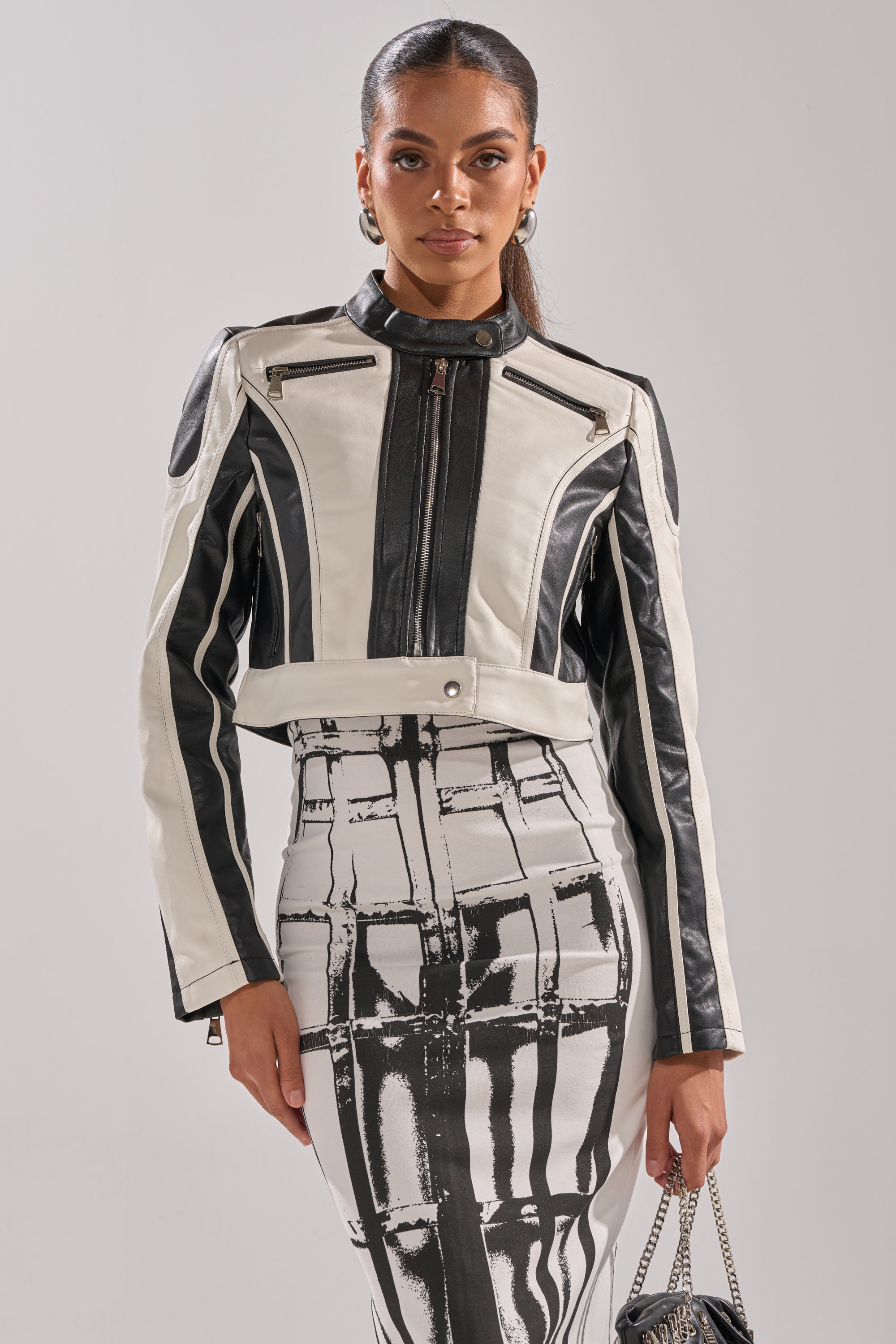 A woman stands confidently in the BETTER LUCK CROPPED MOTO IN BLACK WHITE, a faux leather black and white jacket with zipper details, paired with a matching high-waisted skirt featuring a bold abstract print. 0