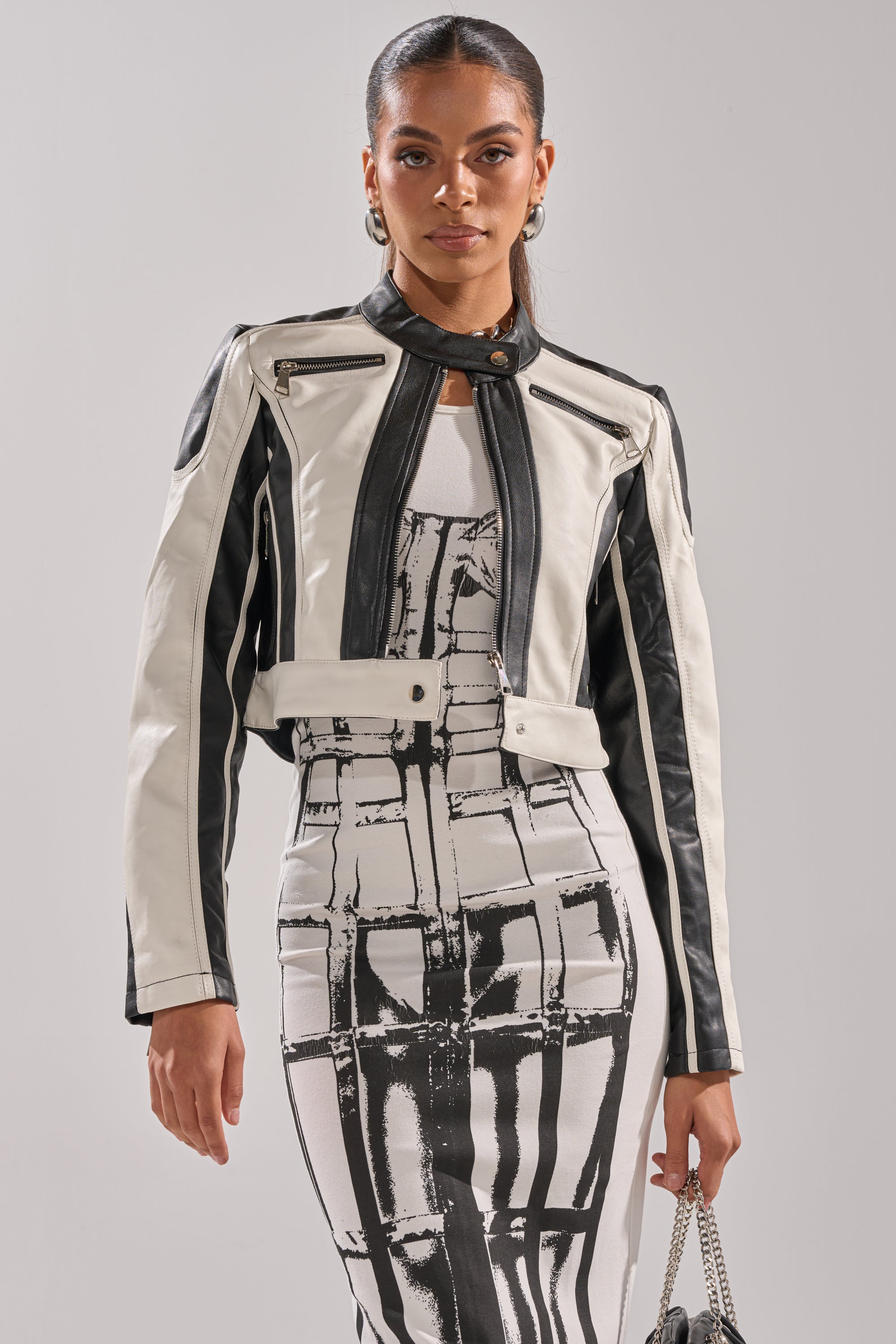 A woman poses confidently against a plain background, wearing the BETTER LUCK CROPPED MOTO IN BLACK WHITE over a fitted patterned dress. She holds a small purse and has her hair pulled back. 1
