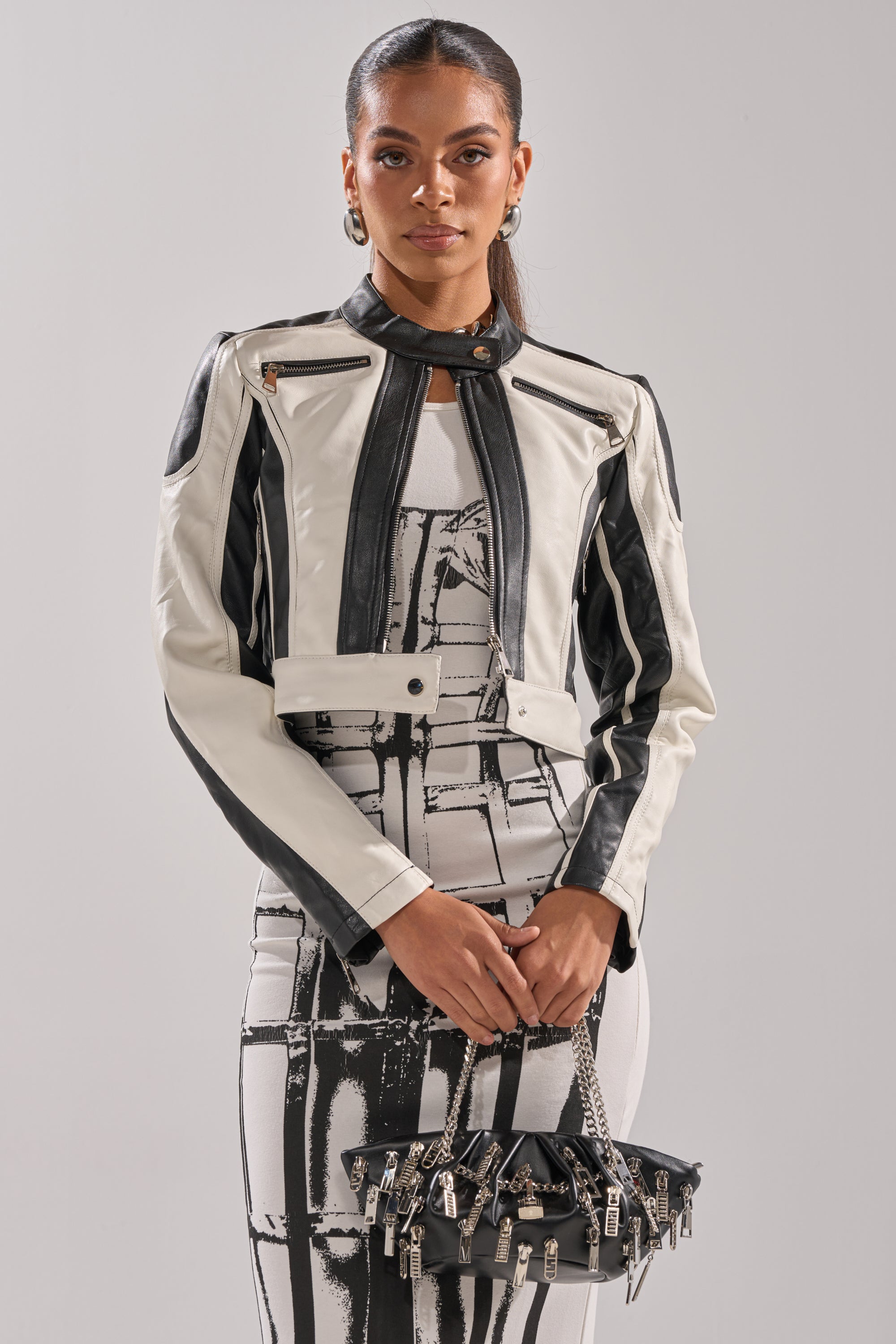 A woman wears the BETTER LUCK CROPPED MOTO IN BLACK WHITE over a patterned dress, holding a small black handbag with metallic details against a plain light background. 2