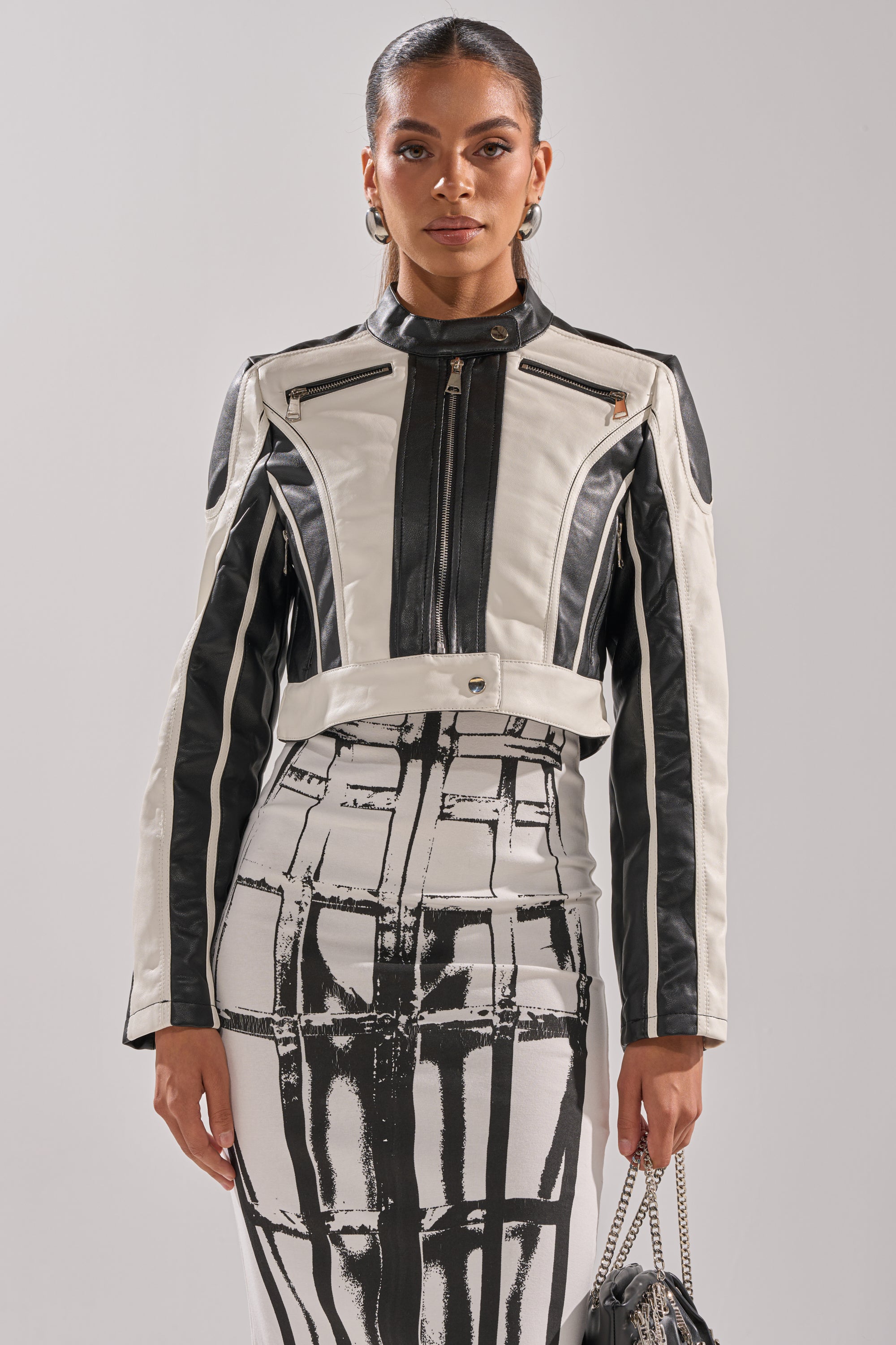 A woman faces forward in the BETTER LUCK CROPPED MOTO IN BLACK WHITE, styled with a matching skirt featuring a bold geometric print and holding a small chain-strap bag against a plain light background. 3