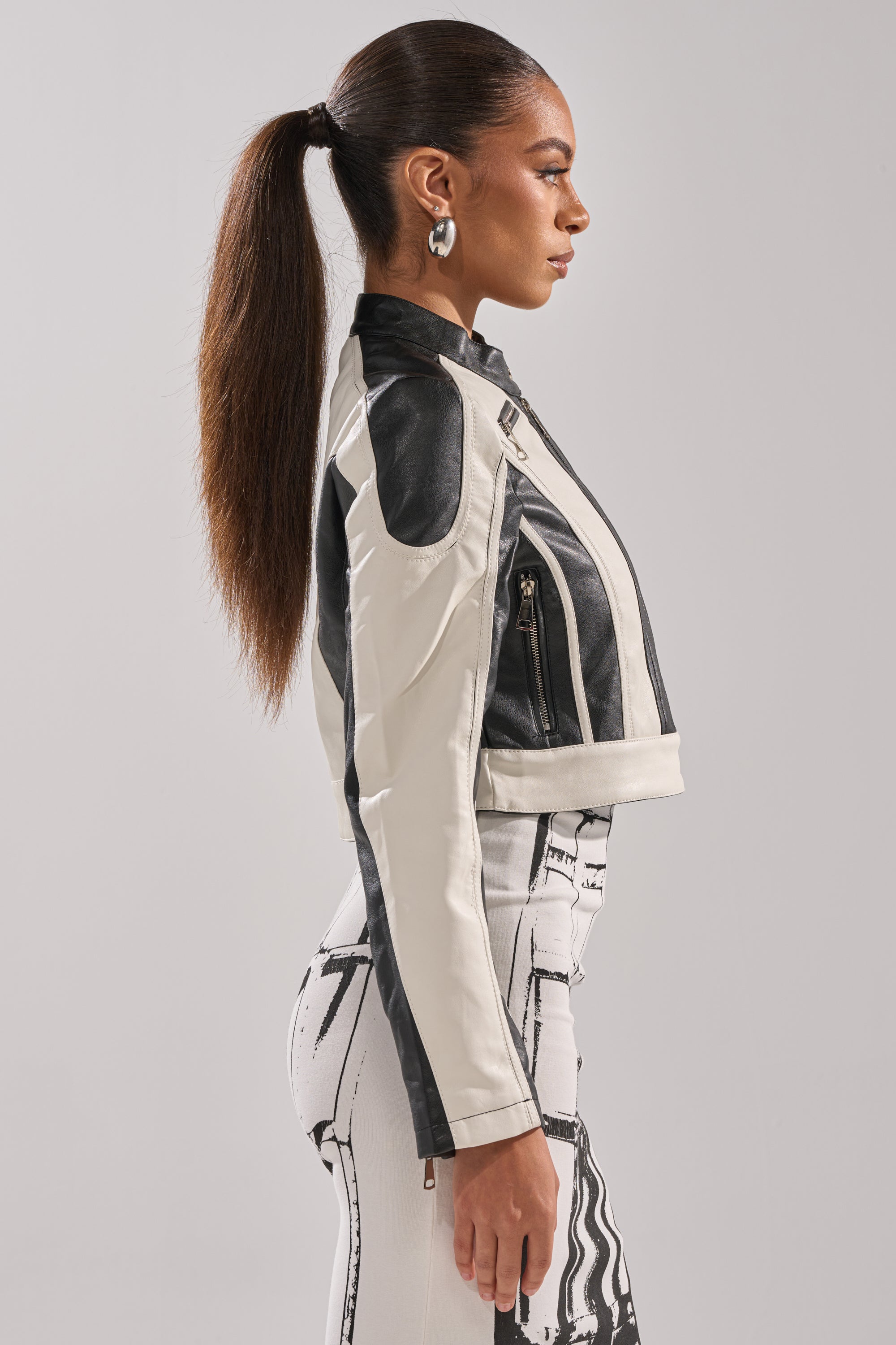 A woman with long, straight hair in a ponytail models the BETTER LUCK CROPPED MOTO IN BLACK WHITE and high-waisted graphic pants, standing in profile against a light gray background. 4