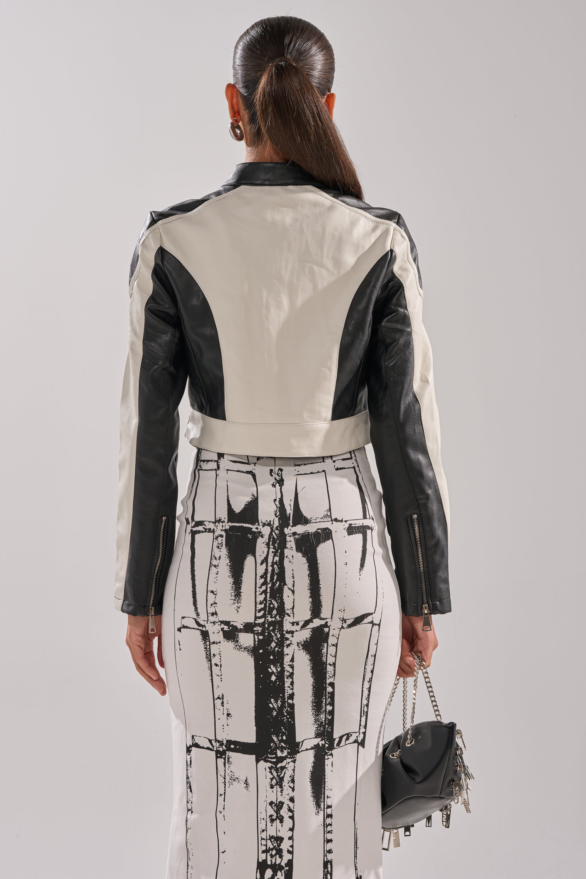 A woman with slicked-back hair is seen from behind wearing the BETTER LUCK CROPPED MOTO IN BLACK WHITE, a high-waisted skirt with a bold graphic print, and holding a small black handbag with silver accents. 5