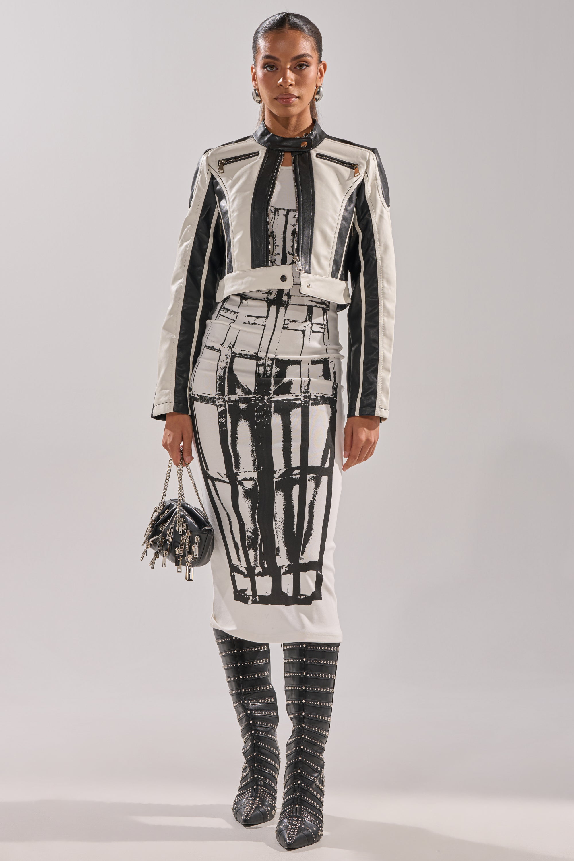 A woman wears the BETTER LUCK CROPPED MOTO IN BLACK WHITE jacket, a matching abstract-patterned knee-length dress, studded knee-high boots, and carries a small black purse with metal details. 6